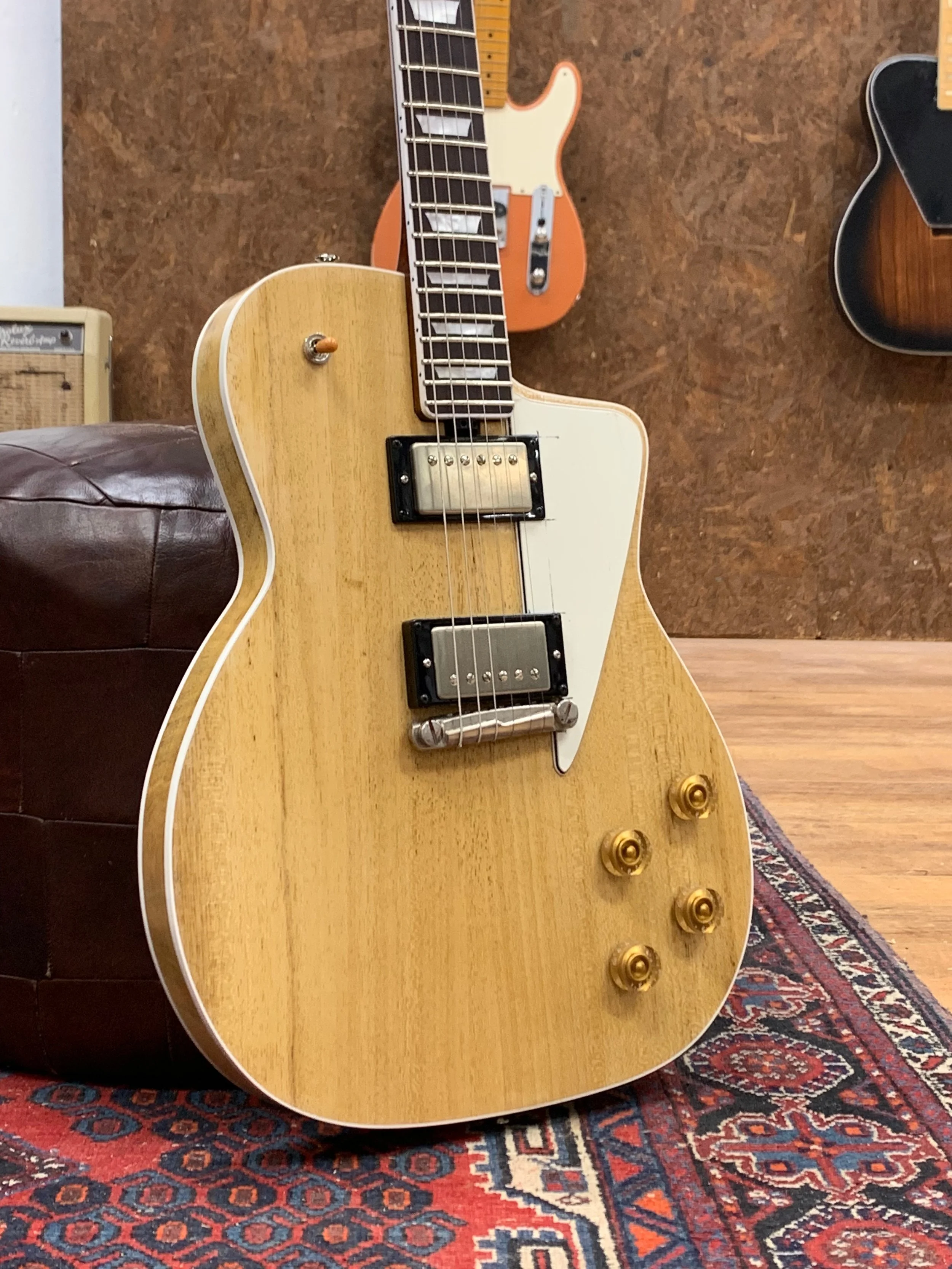 Een elektrische gitaar met een natuurlijke houtafwerking ligt op een tapijt. Op de achtergrond zijn andere gitaren zichtbaar.