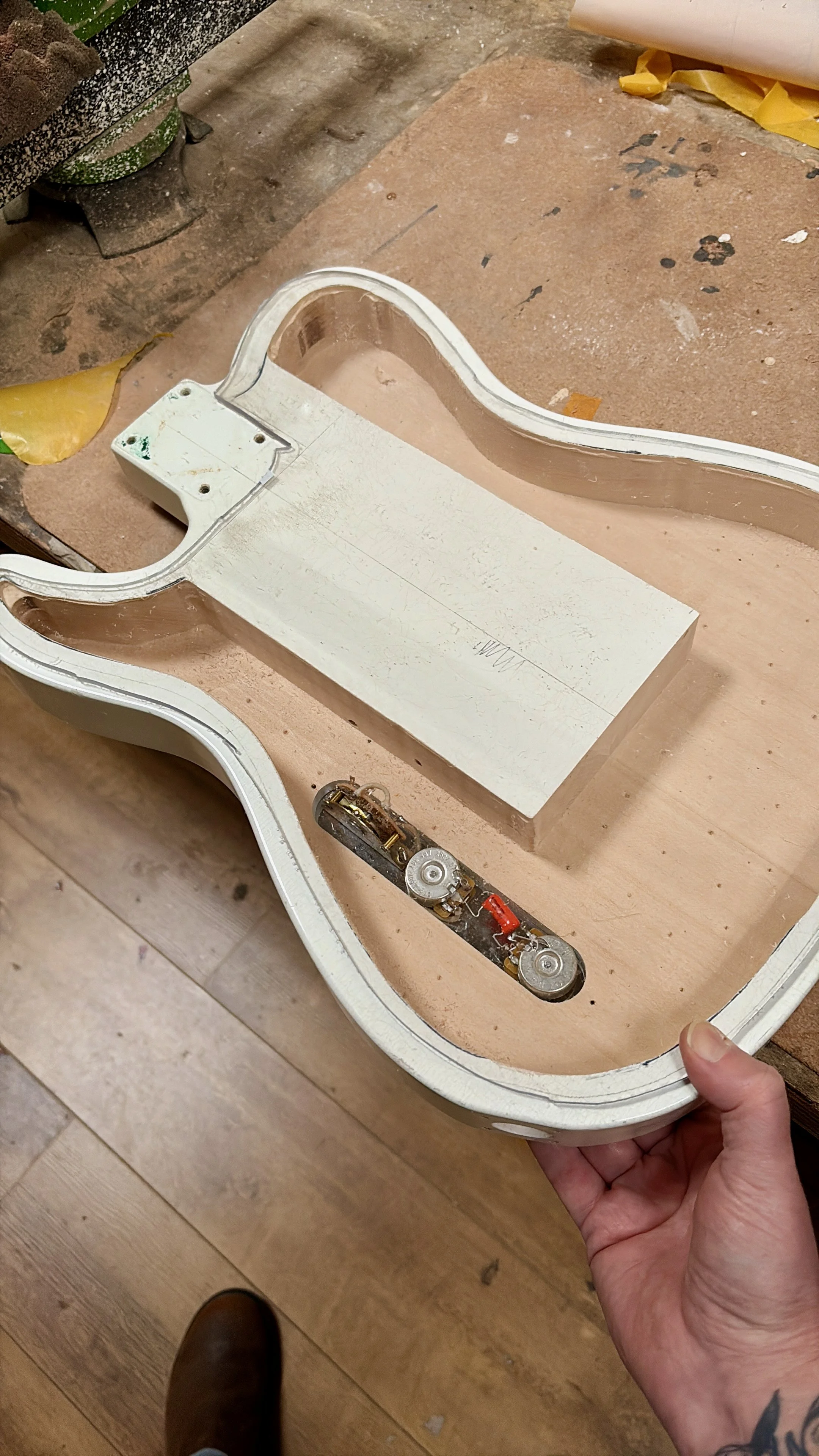 Een hand houdt een witte, gelijmde gitaarkast met een interne mechanische component, op een werkbank met gereedschap en verfspatten.