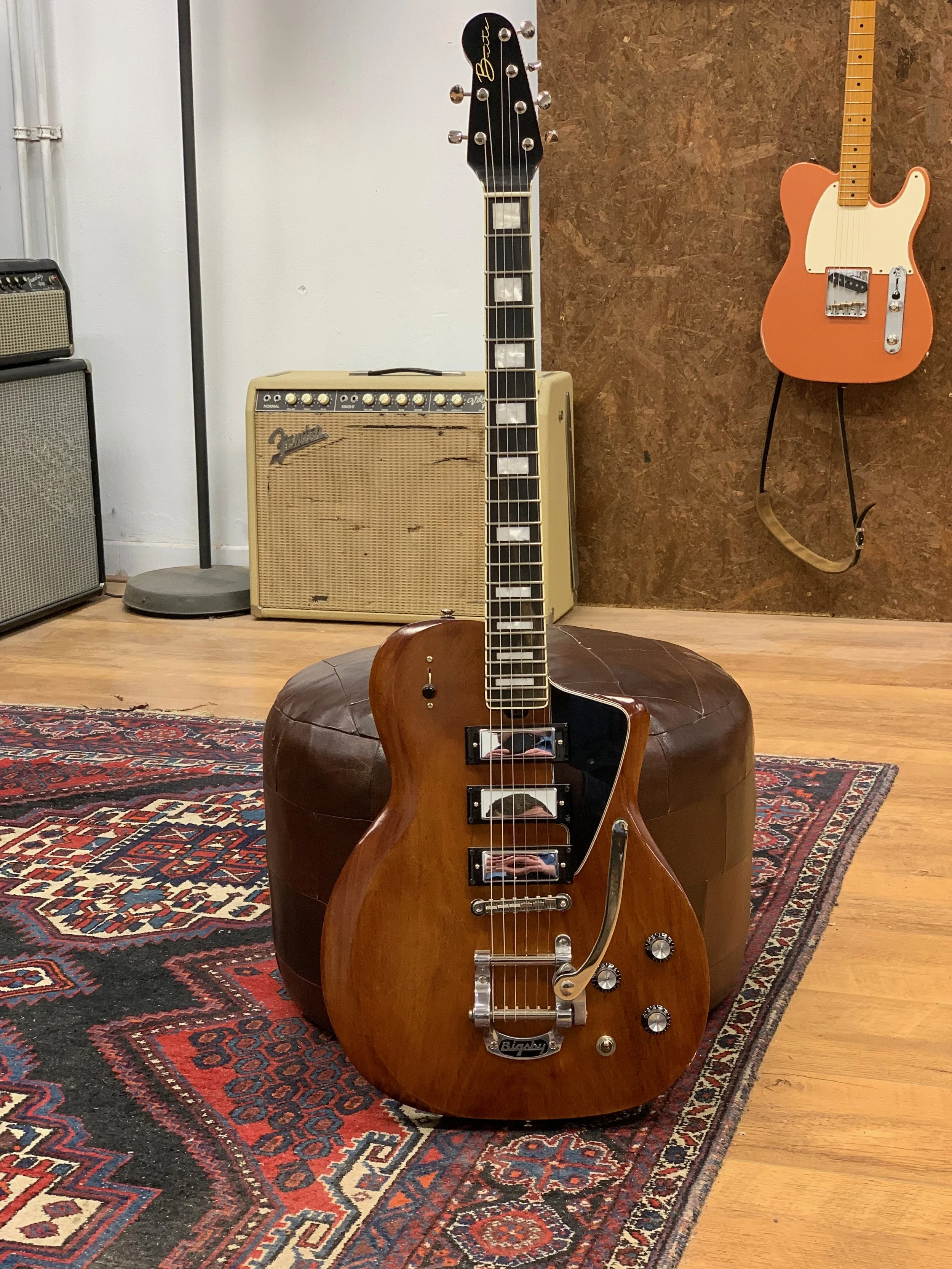 Een elektrische gitaar met een houten body staat op een ronde poef, op een tapijt. Op de achtergrond hangen een versterker en een elektrische gitaar aan de muur.