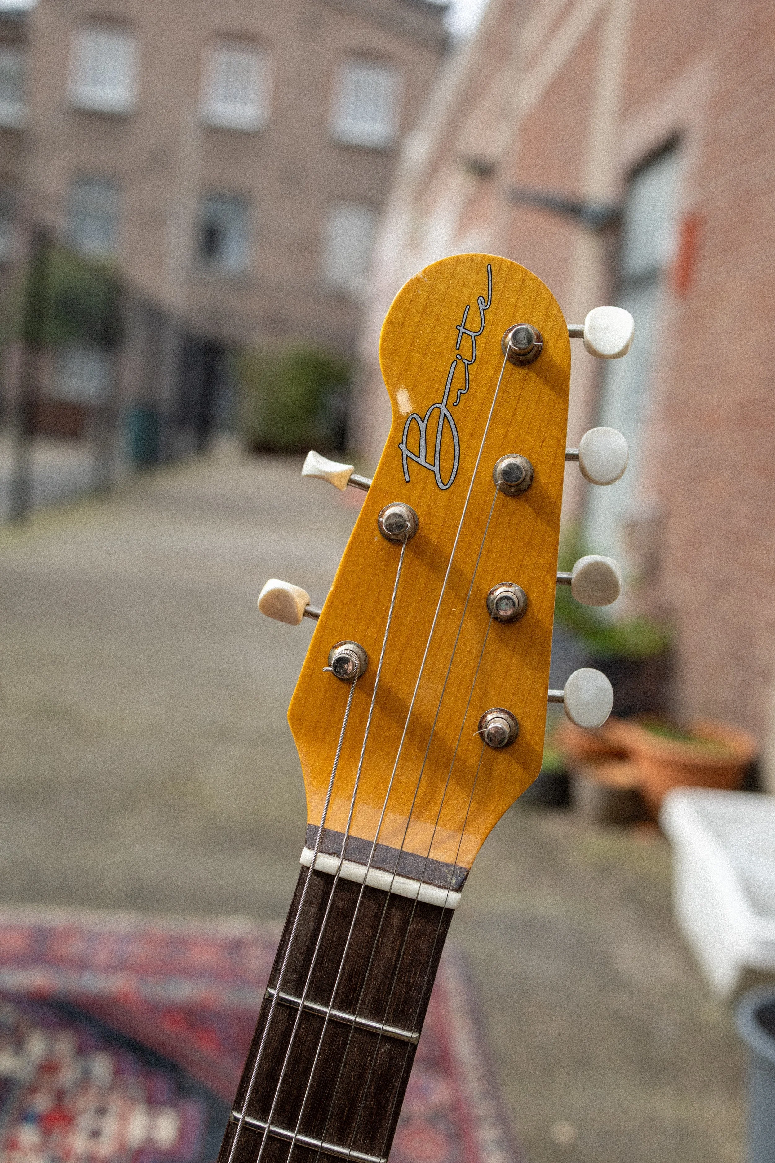 De afbeelding toont het hoofd van een gitaar met de naam 'Arteci' geschreven op de kop. De kop bevat zes mechanische stemknoppen en snaren, met een achtergrond van een onduidelijke binnenruimte.