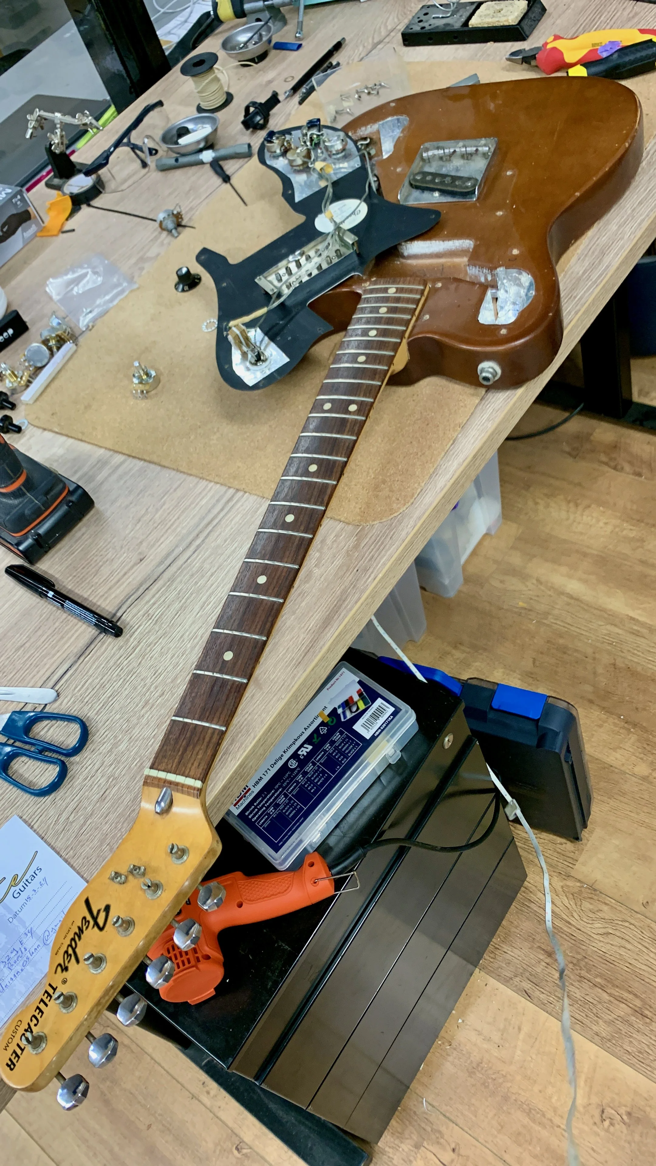 Een elektrische gitaar wordt gerepareerd of opgebouwd op een werkbank, met verschillende gereedschappen en onderdelen rondom.