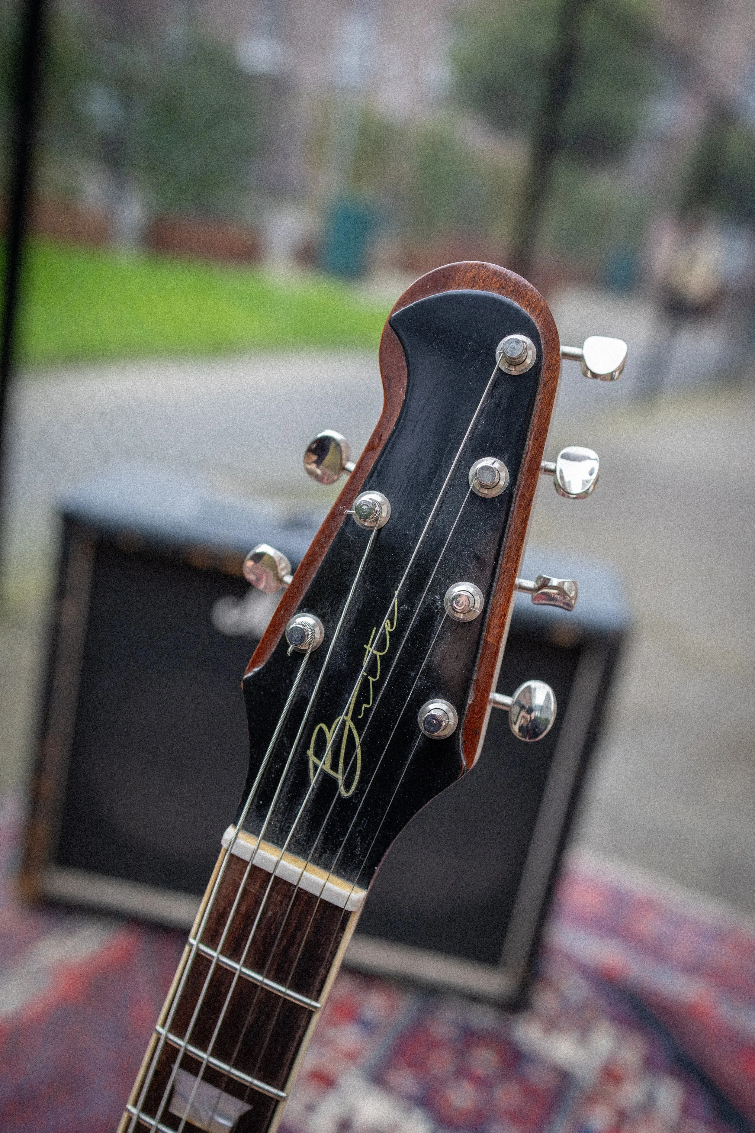 Close-up van een gitaarkop met tuning pegs en snaren, met een blur achtergrond van een kamer en een tapijt.