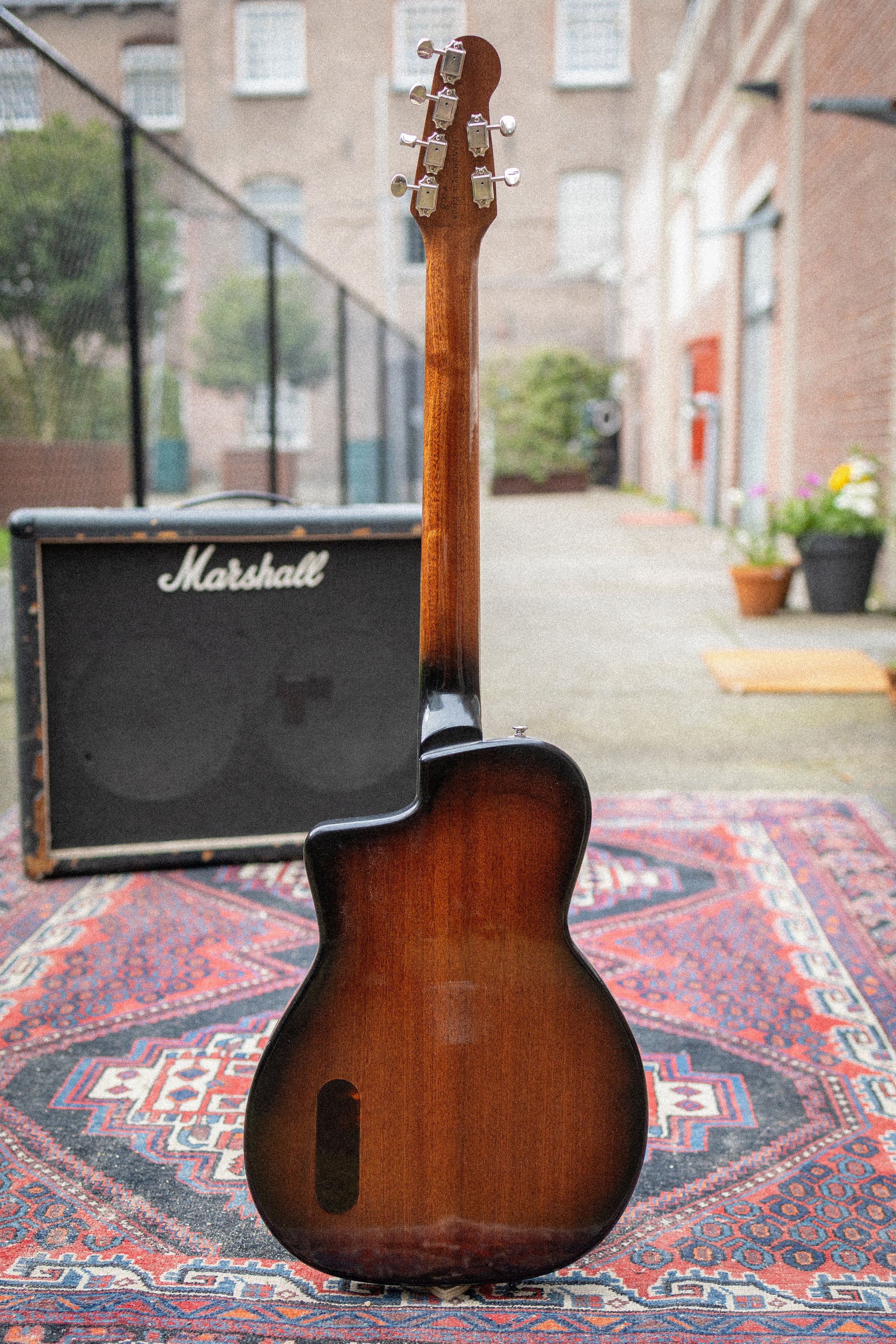 Een houten elektrische gitaar staat op een traditioneel tapijt. Op de achtergrond zijn een Marshall versterker en buitenrondingen te zien, waaronder een tuin met planten en een huismuur.