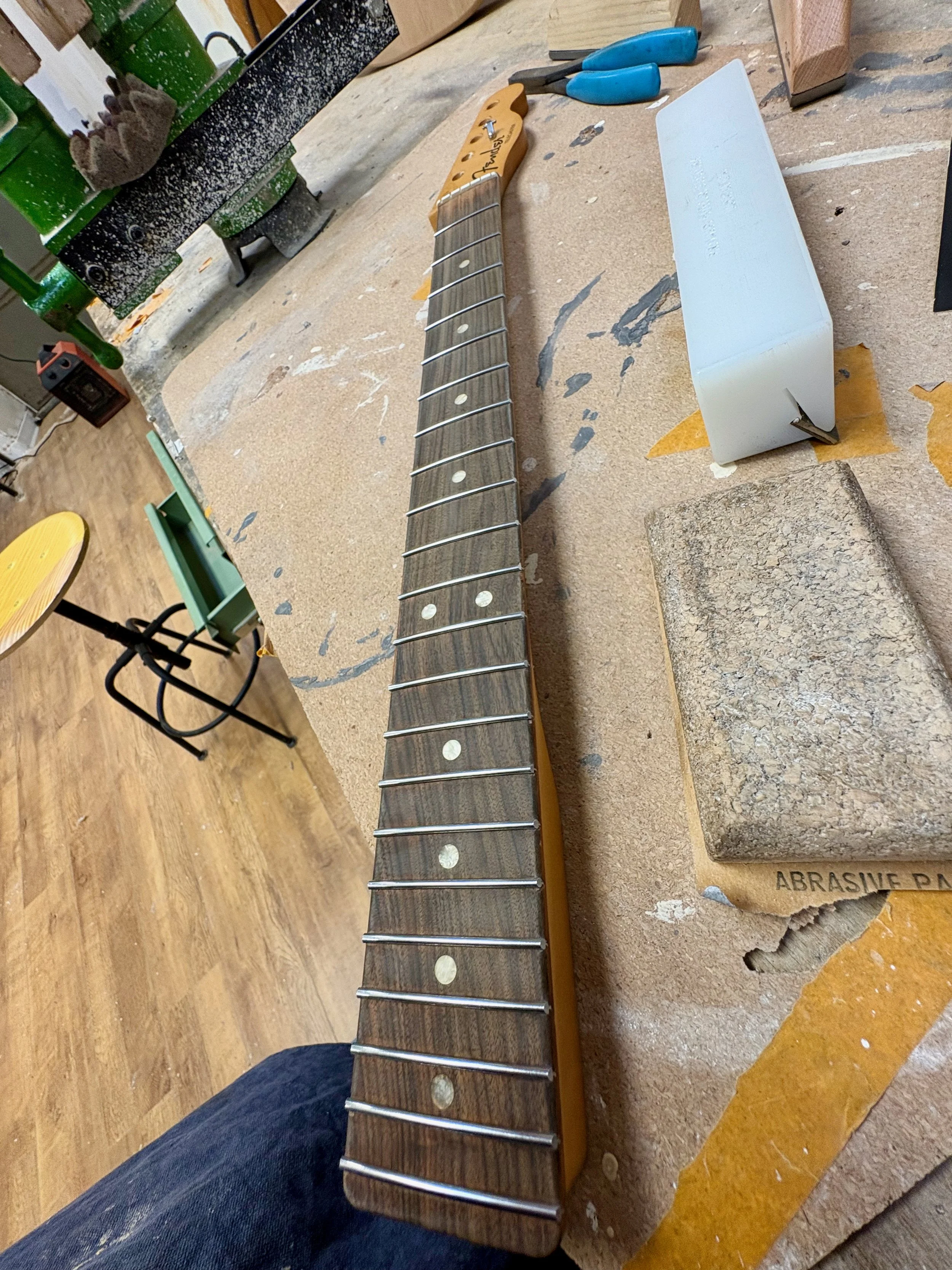 Een werkbank met gitaarhals, schuurblok, schroevendraaier, handschoenen en andere gereedschappen in een werkplaats.