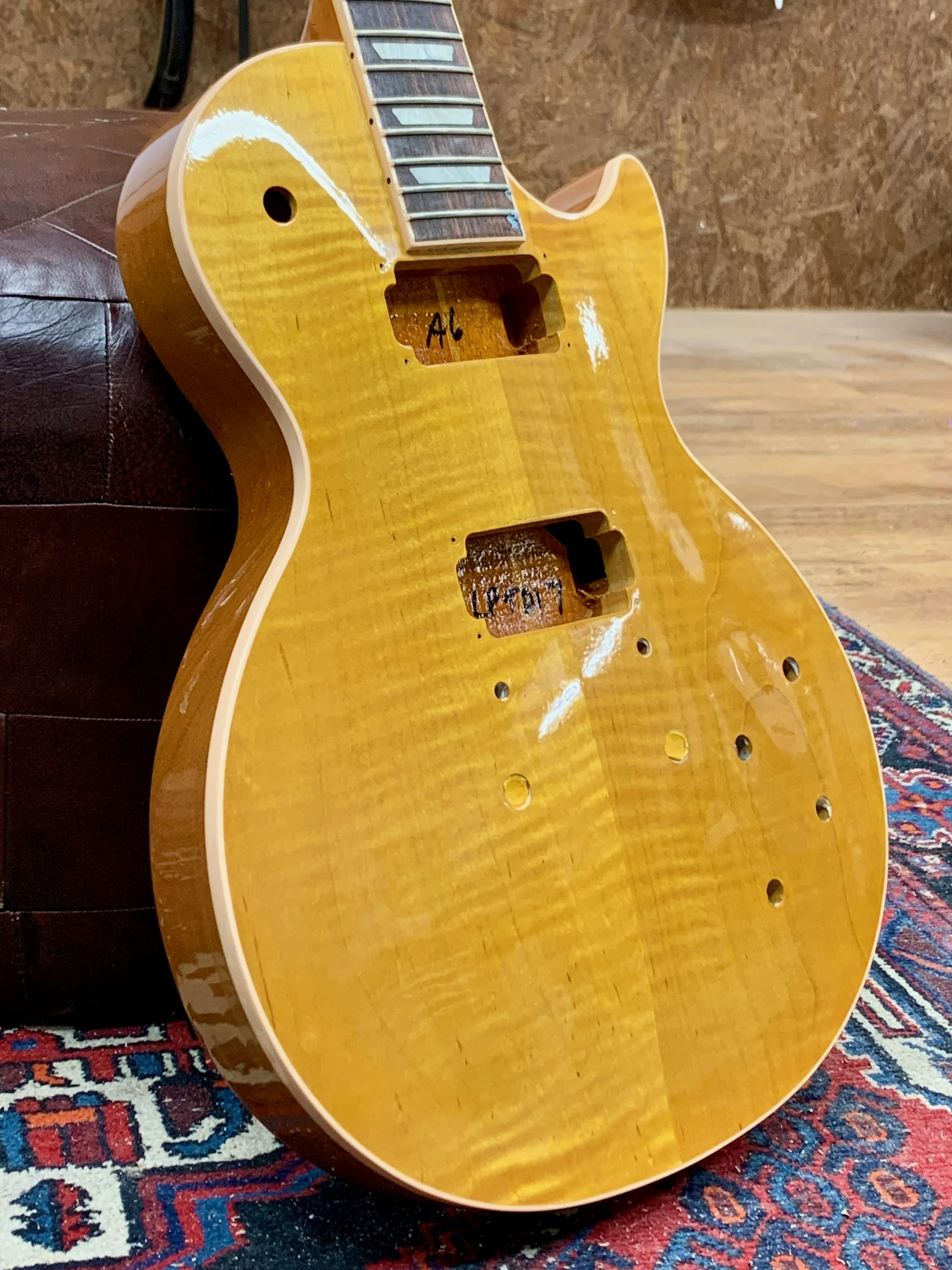 De afbeelding toont de body van een ongeassembleerde elektrische gitaar, liggend op een tapijt en tegen een muur, zonder snaren en hardware. Op de gitaar is een inscriptie te zien met de tekst 'AL' en 'Cech'.