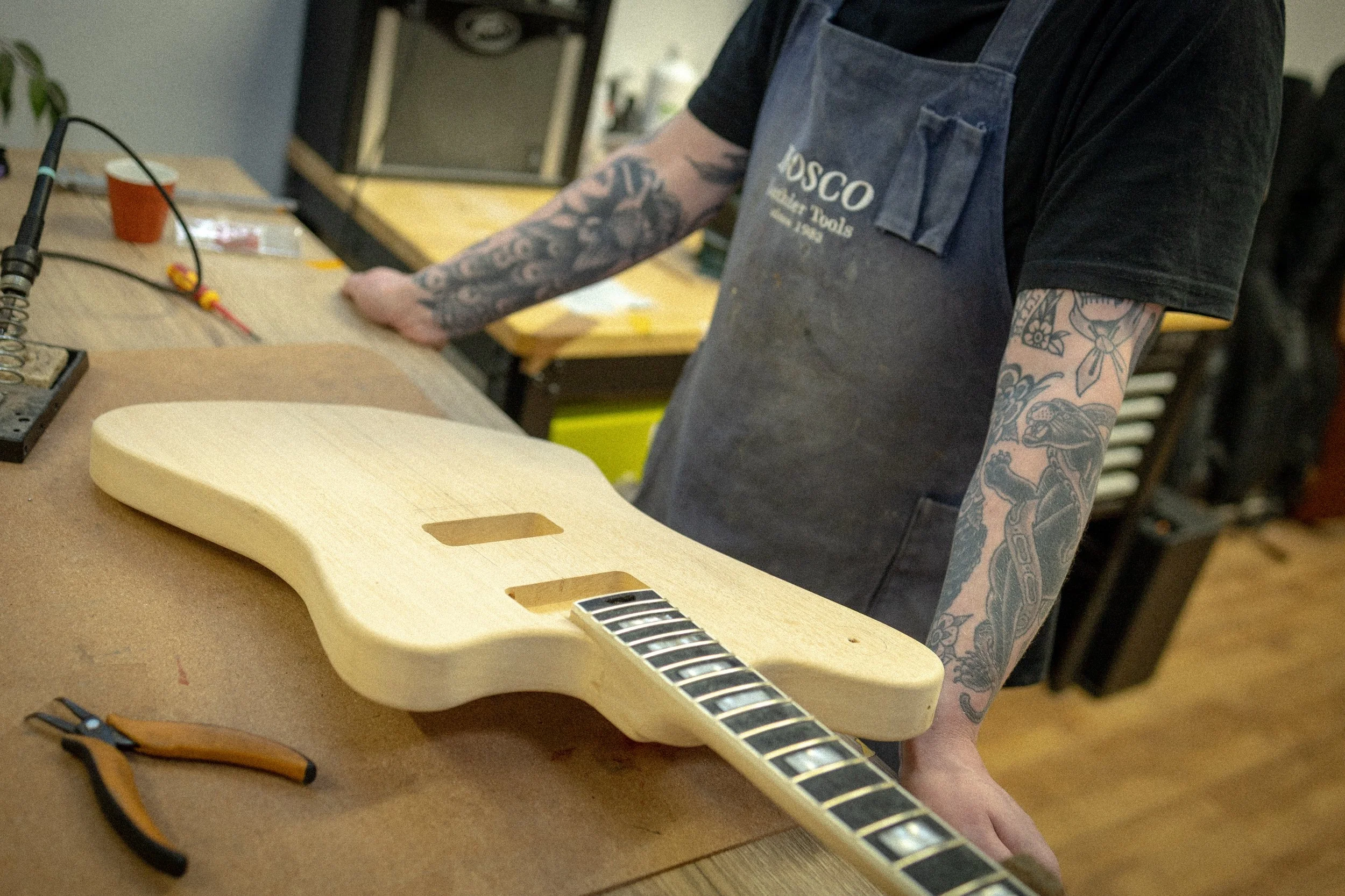 Een persoon met getatoeëerde armen werkt aan de montage van een elektrische gitaar in een werkplaats. De gitaar wordt voorbereid en de persoon gebruik tools op een werkbank.
