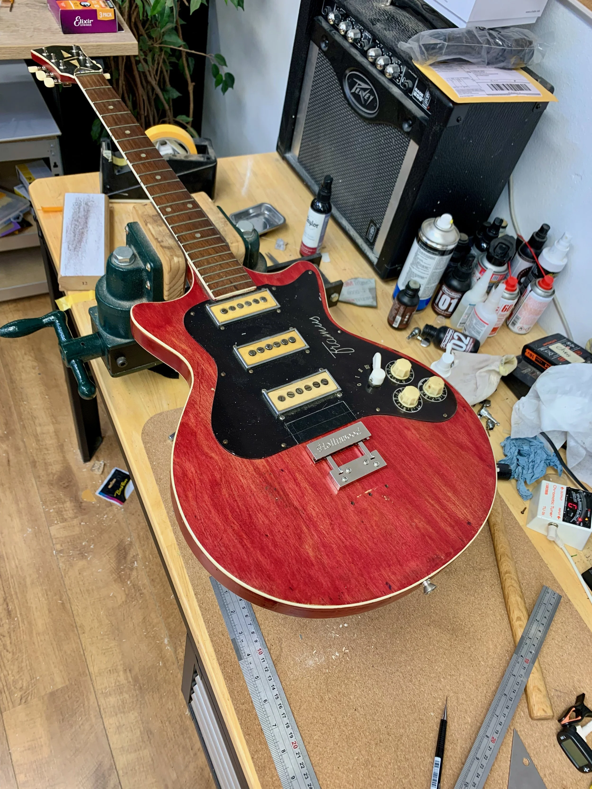 Een werktafel met een rode elektrische gitaar in reparatie, omringd door meetlinten, gereedschap en reparatiespullen. Op de achtergrond staan een kleine versterker, schroevendraaier, verf en lakken, en andere gereedschappen en materialen voor het her