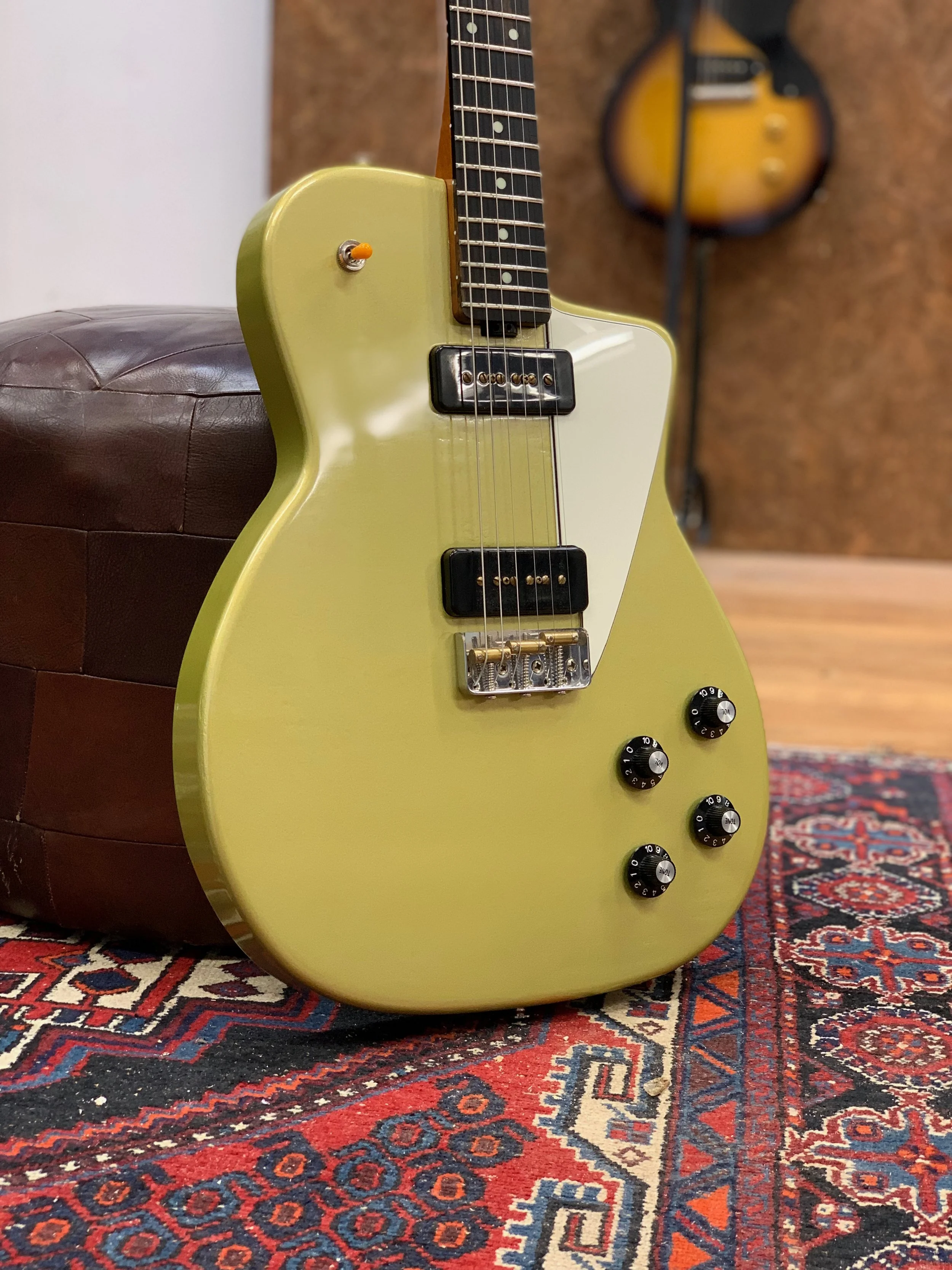 Geel elektrische gitaar met zwarte hals en vier controleknoppen op een houten vloer met een rood, blauw en beige geruite tapijt.