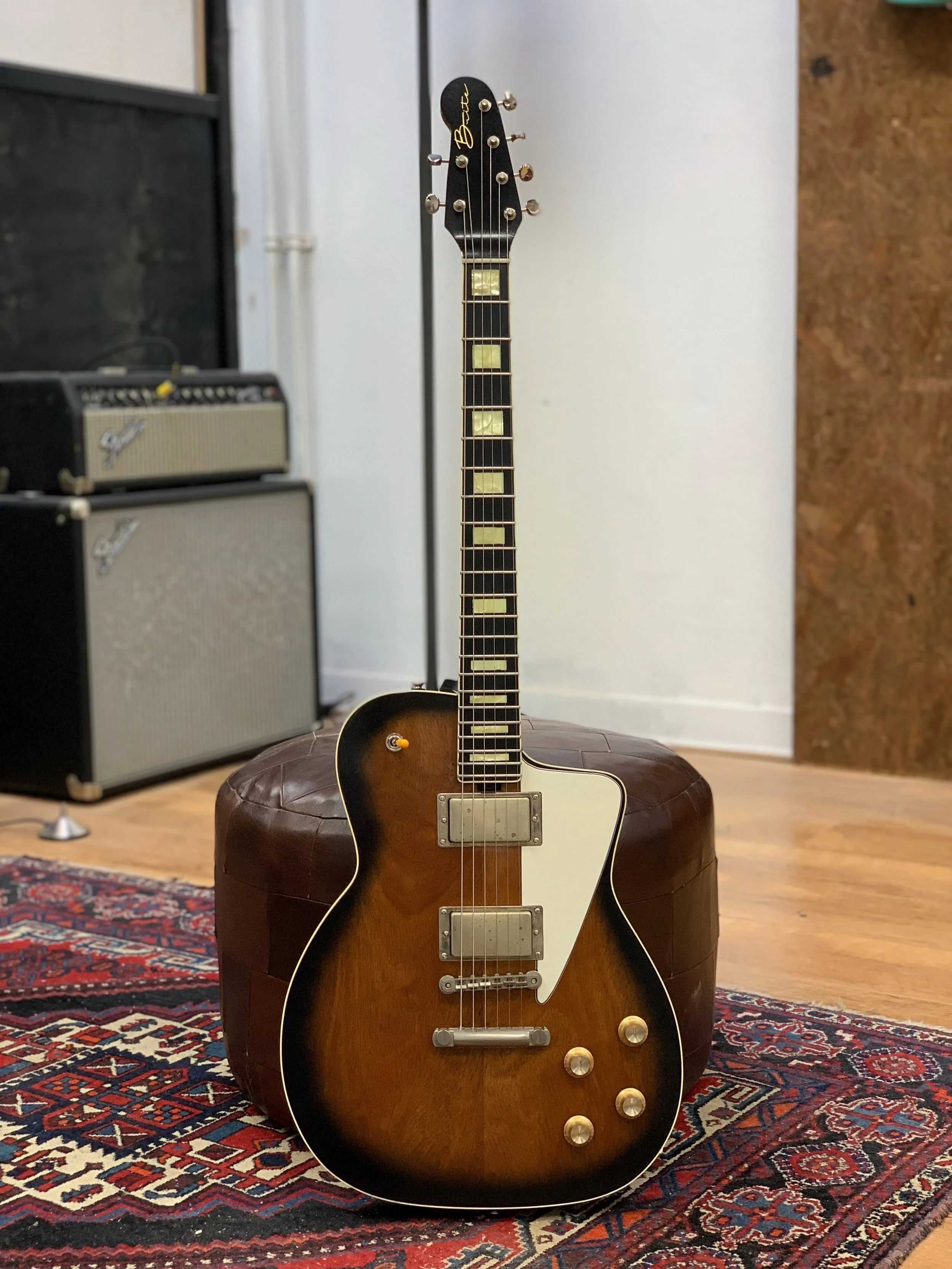 Een vintage elektrische gitaar met een bruine houten body en een witte slagplaat, geplaatst op een ronde, donkerbruine opblaasbare poef op een traditionele tapijt. Op de achtergrond zijn twee kleine zwart-witte versterkers te zien.
