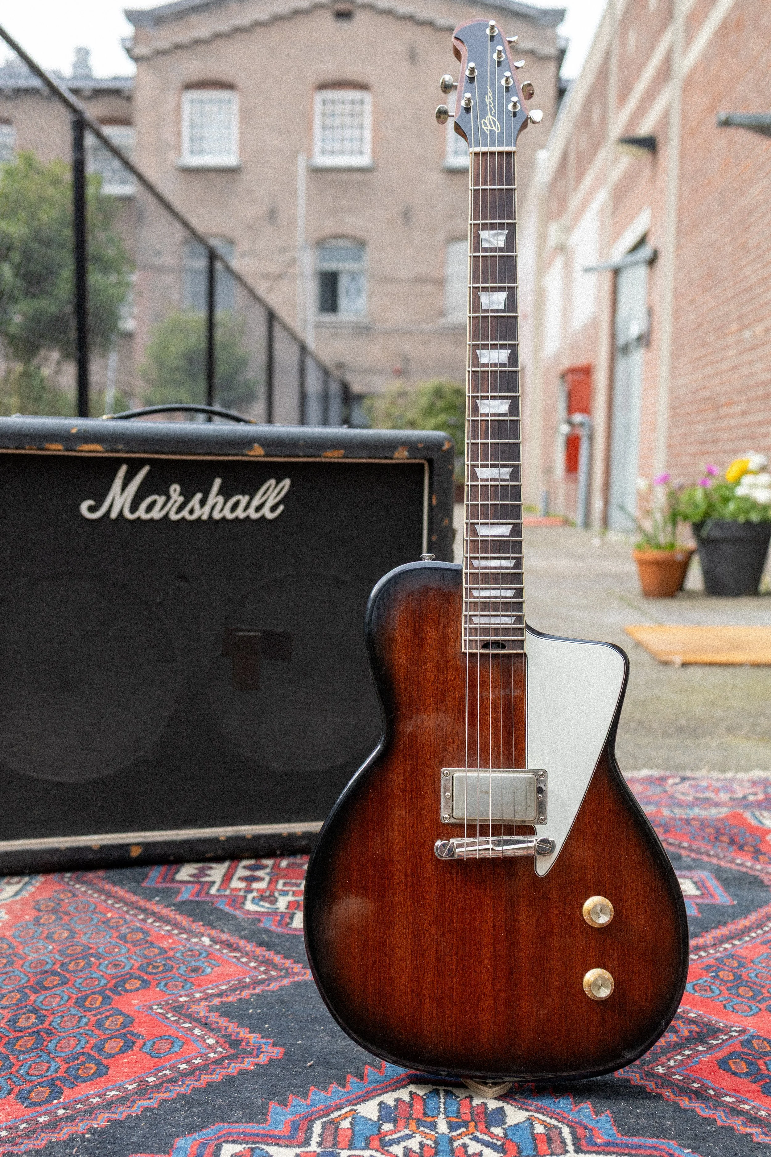 Een elektrische gitaar met een bruine houtkleur en wit mozaïek inleg op de hals, geplaatst op een kleurrijk tapijt, met een Marshall versterker op de achtergrond, buiten bij een gebouw met bakstenen muren en een tuin met bloemen.