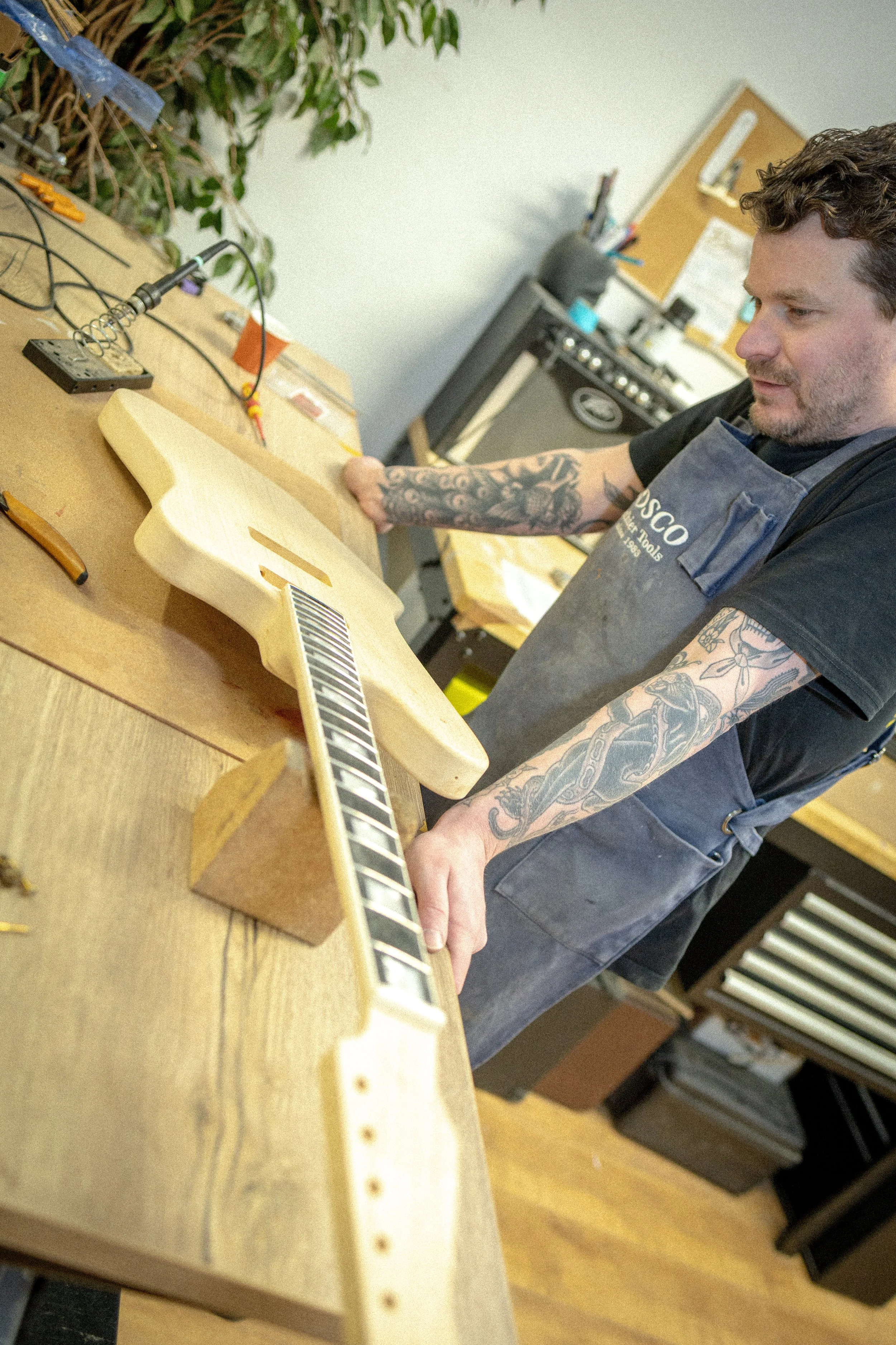 Man met tattoos op armen werkt aan een gitaar in een werkplaats, omringd door gereedschap en apparatuur.