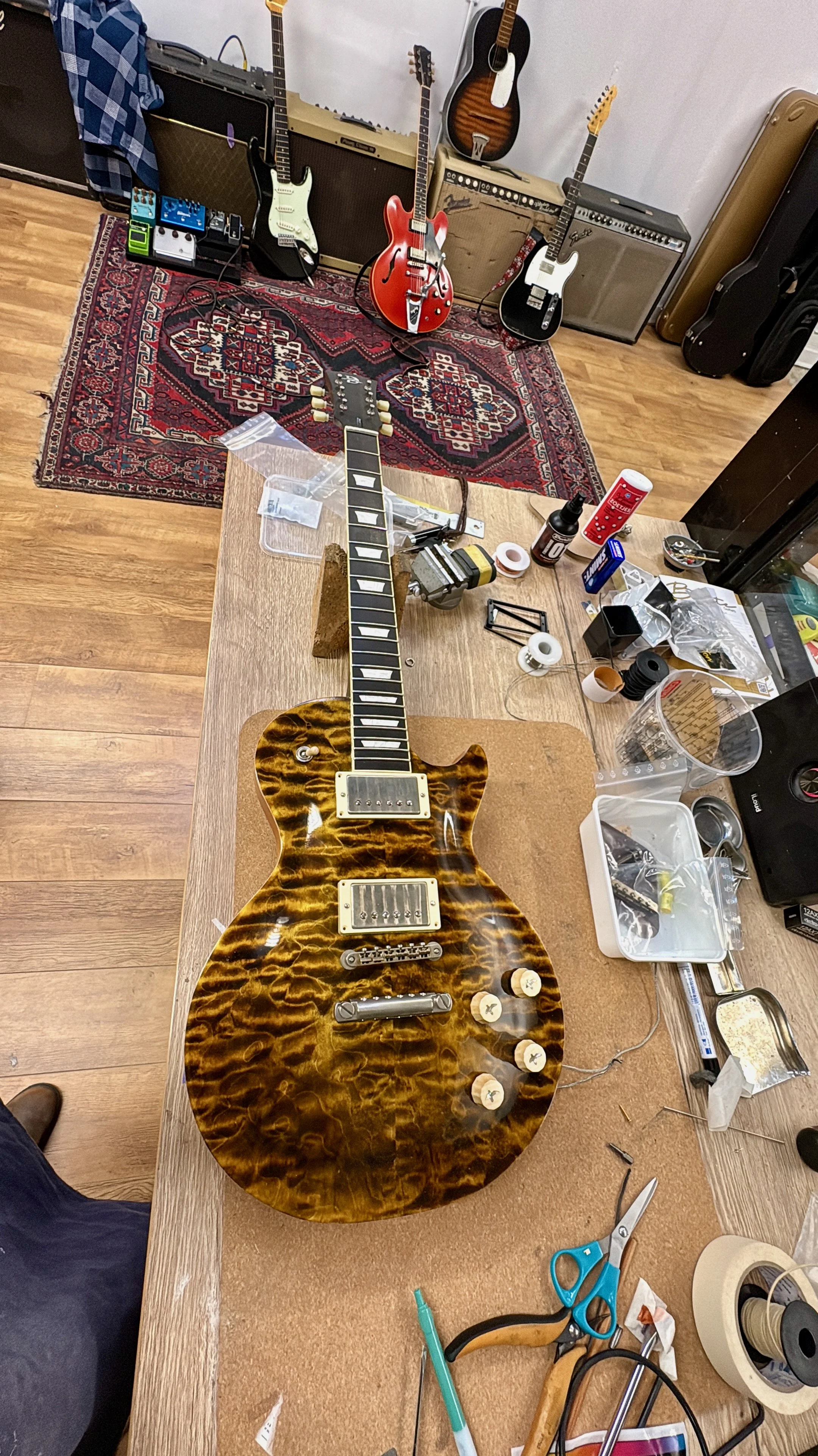 Een werkbank met een ongeslepen elektrische gitaar met een bắtterybruine, getwiste afwerking. Op de werkbank liggen gereedschap en onderdelen zoals scharen, een soldeerblam, schroeven en tape, om de gitaar te repareren of te assembleren.