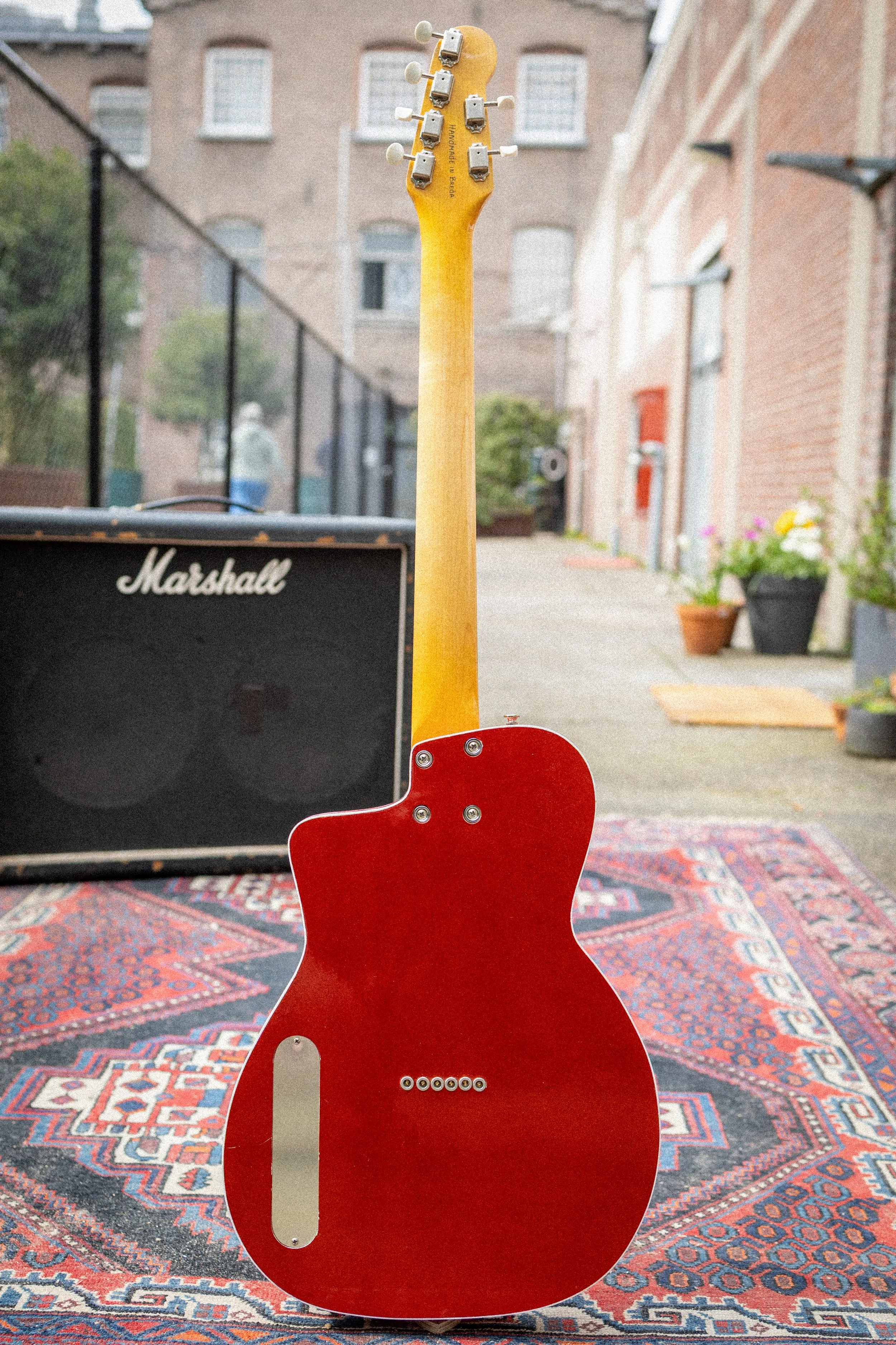 een rode gitaar met jamplek op een kleurrijk tapijt, met een zwart versterker links en een background van een stenen gebouw met ramen en bloempotten.