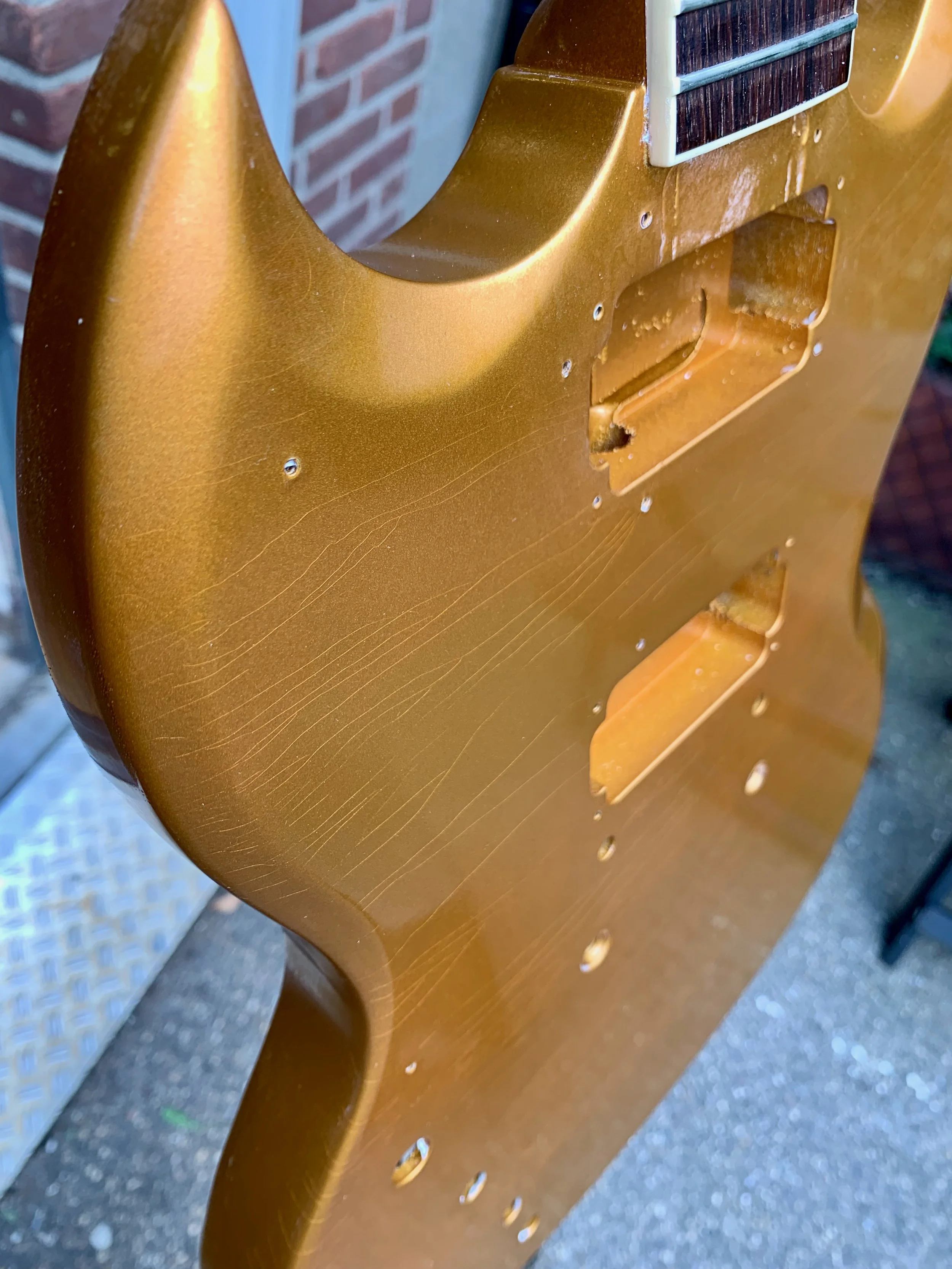 De afbeelding toont een dicht bij een bruin-gouden elektrische gitaar, die zich op een buitenlocatie met een bakstenen muur op de achtergrond bevindt. Het lichaam van de gitaar is zichtbaar, vooral de zijkant met kleine bevestigingsgaten en een uitsp