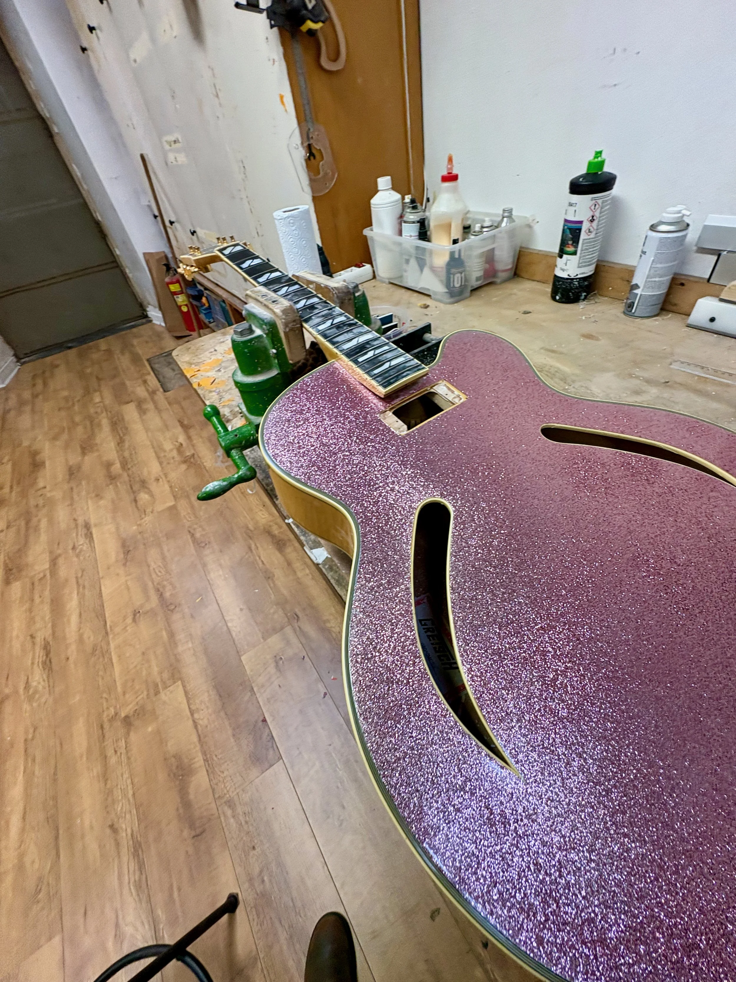 Een hal met een houten vloer en een deur. Op een tafel ligt een beschadigde akoestische gitaar met een paar snaren. Op de achtergrond staan verschillende spullen, inclusief schoonmaakmiddelen en een doos met tools.