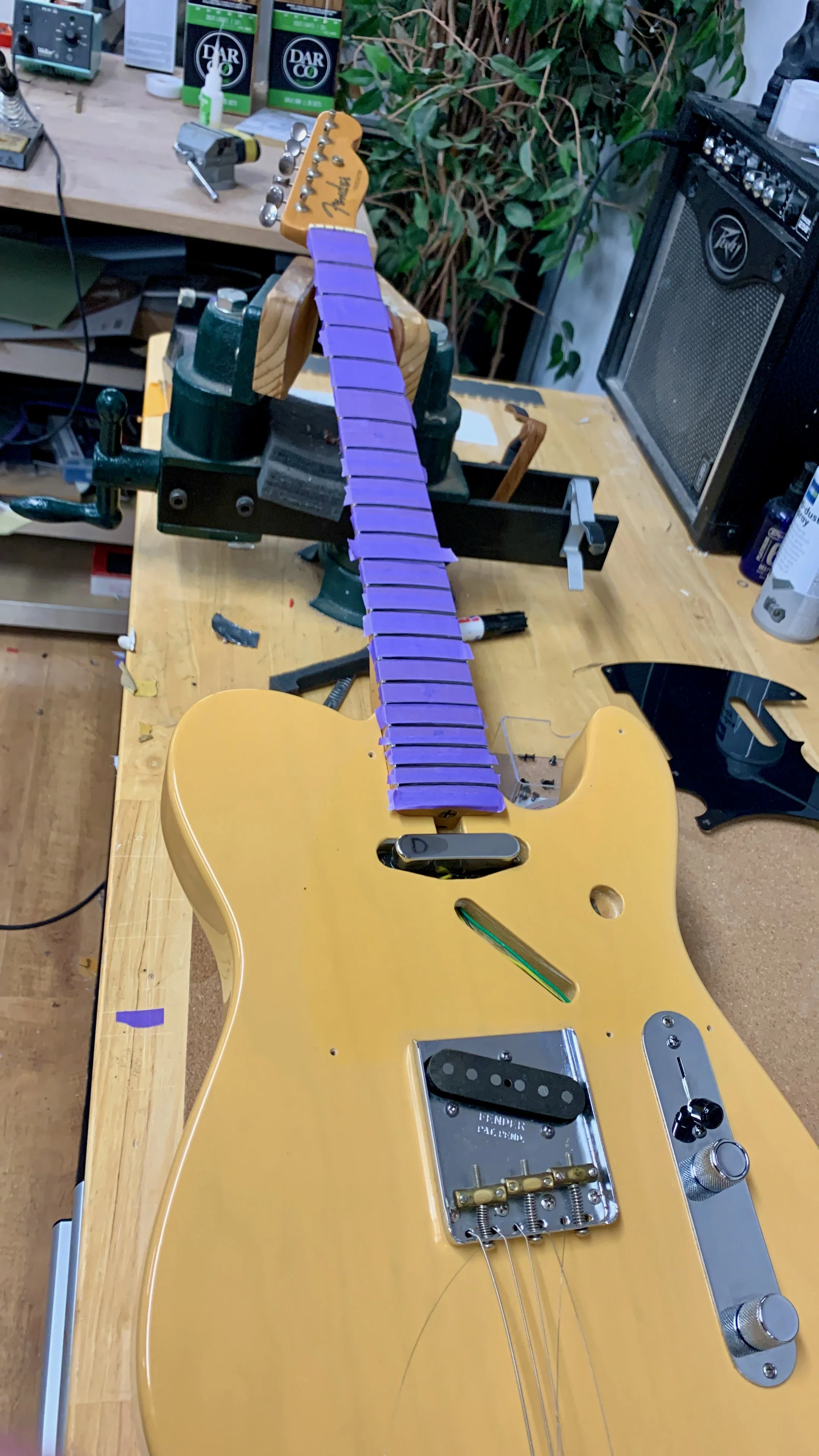 Een gele elektrische gitaar in aanbouw op een werkbank met verschillende gereedschappen en onderdelen, inclusief een uitschuifbare hals en elektronische onderdelen.