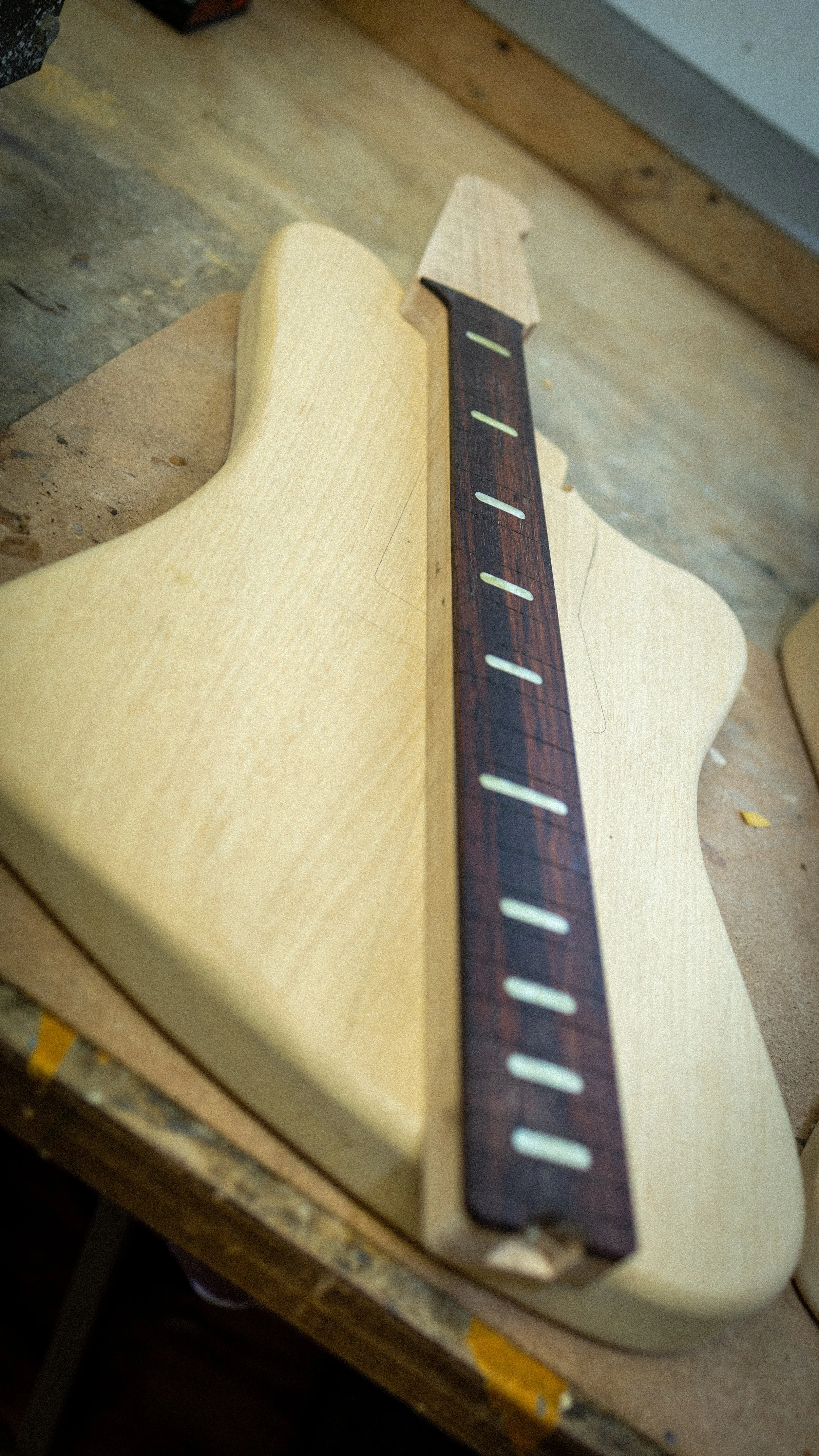 Een gitaarhals ligt op een stuk hout op een werkbank met gereedschap en houtspaanders op de vloer.