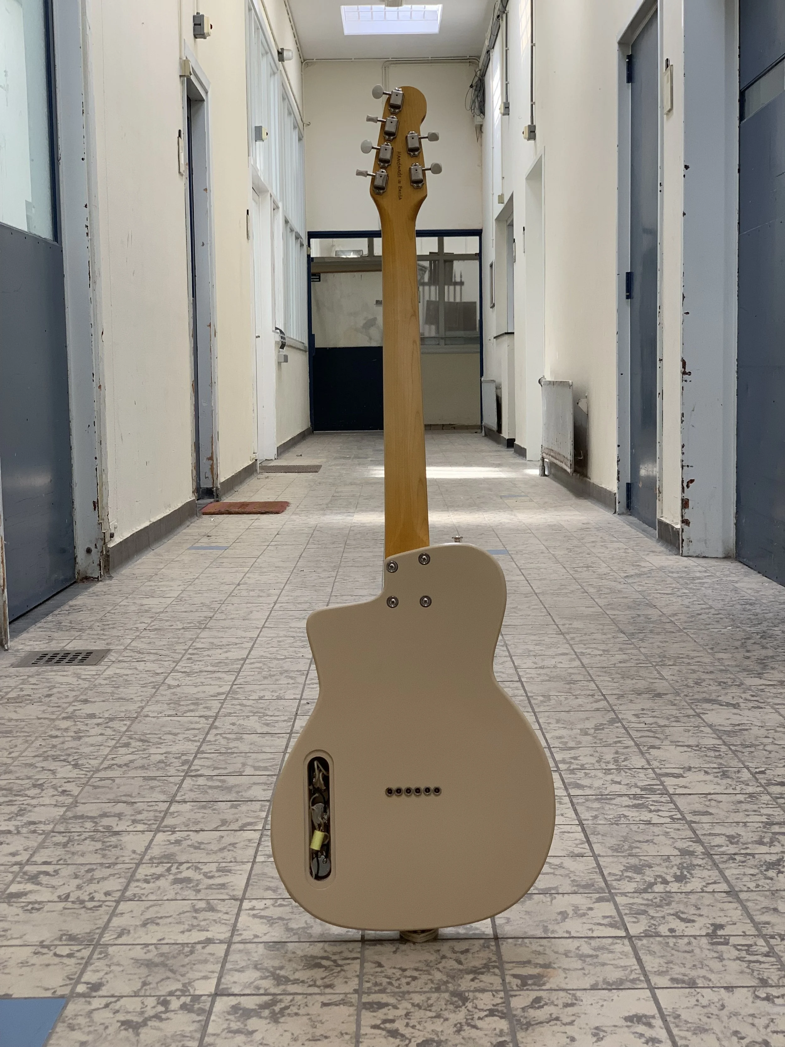 De achterkant van een gitaar wordt gezien, geplaatst op de vloer in een hall met tegels. De gitaar mist de body en toont de binnenkant met draden en mechanieken.