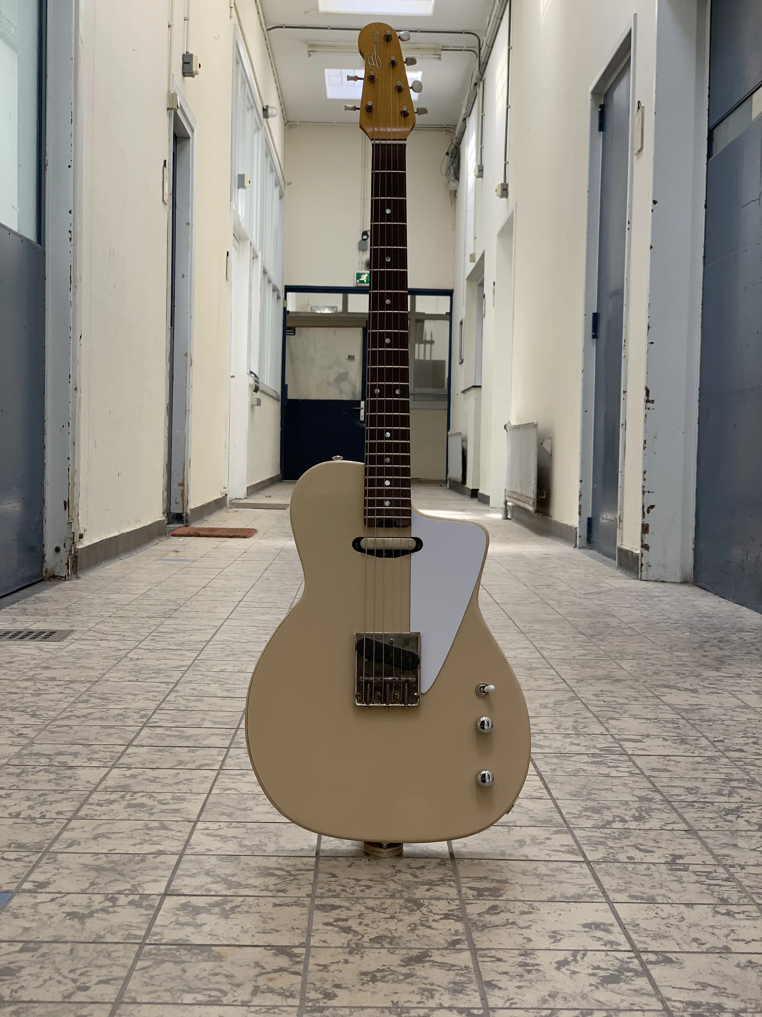 Een ongebruikte blauwe elektrische gitaar met een beige body staat rechtop in een gang met betegelde vloer en panelen muren.