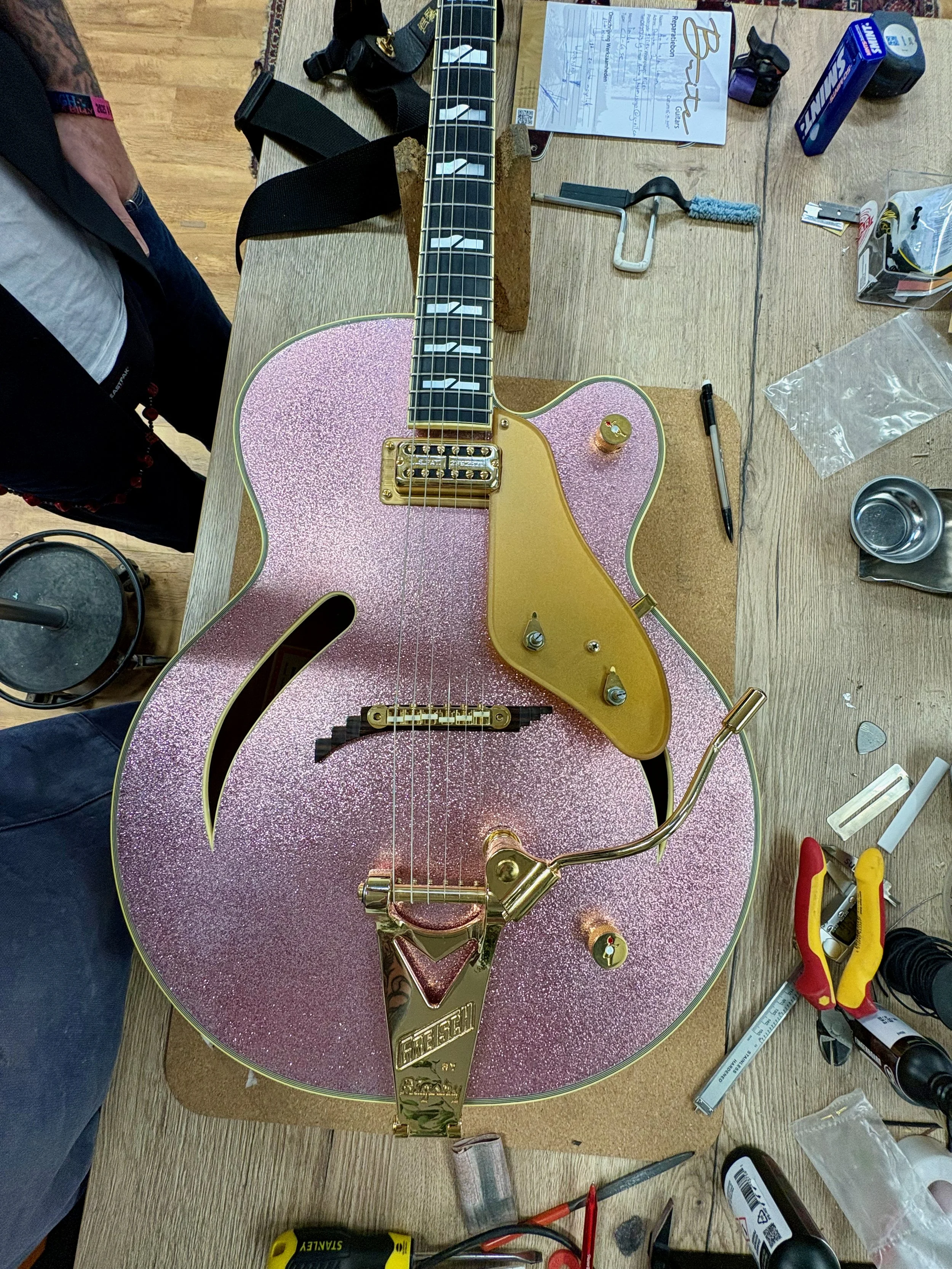 Een glitterende roze gitaar met goudkleurige hardware, drukknoppen en een goudkleurige brug, gelegen op een werkbank omringd door gereedschap en onderdelen.