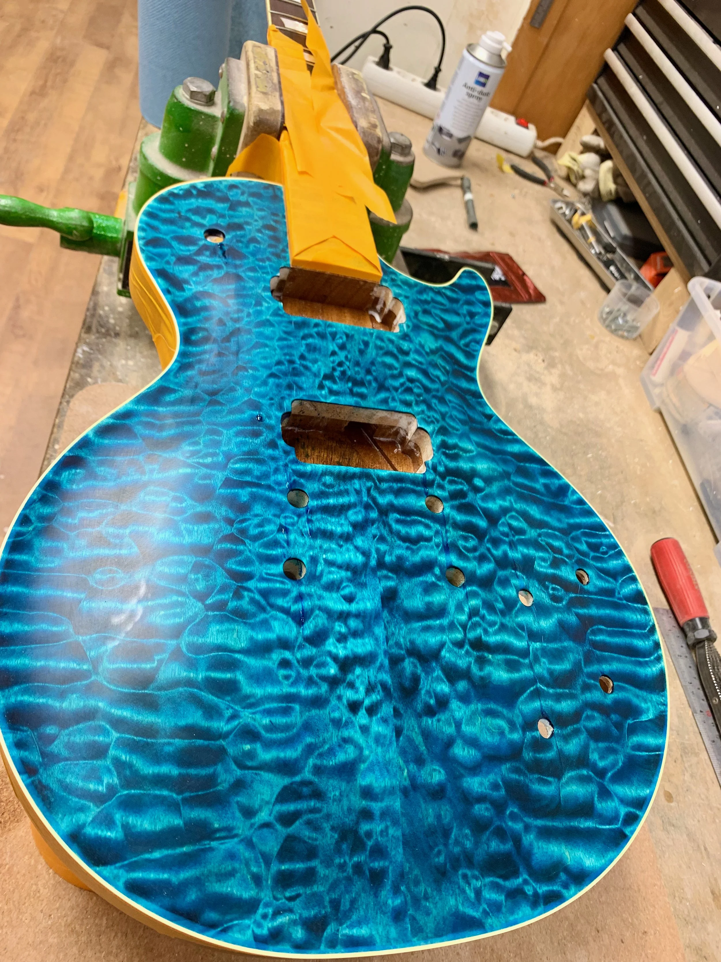 Een elektrische gitaar met een blauwe, getextureerde voorkant ligt op een werkbank in een werkplaats. Er zijn verschillende gereedschappen en materialen op de achtergrond.