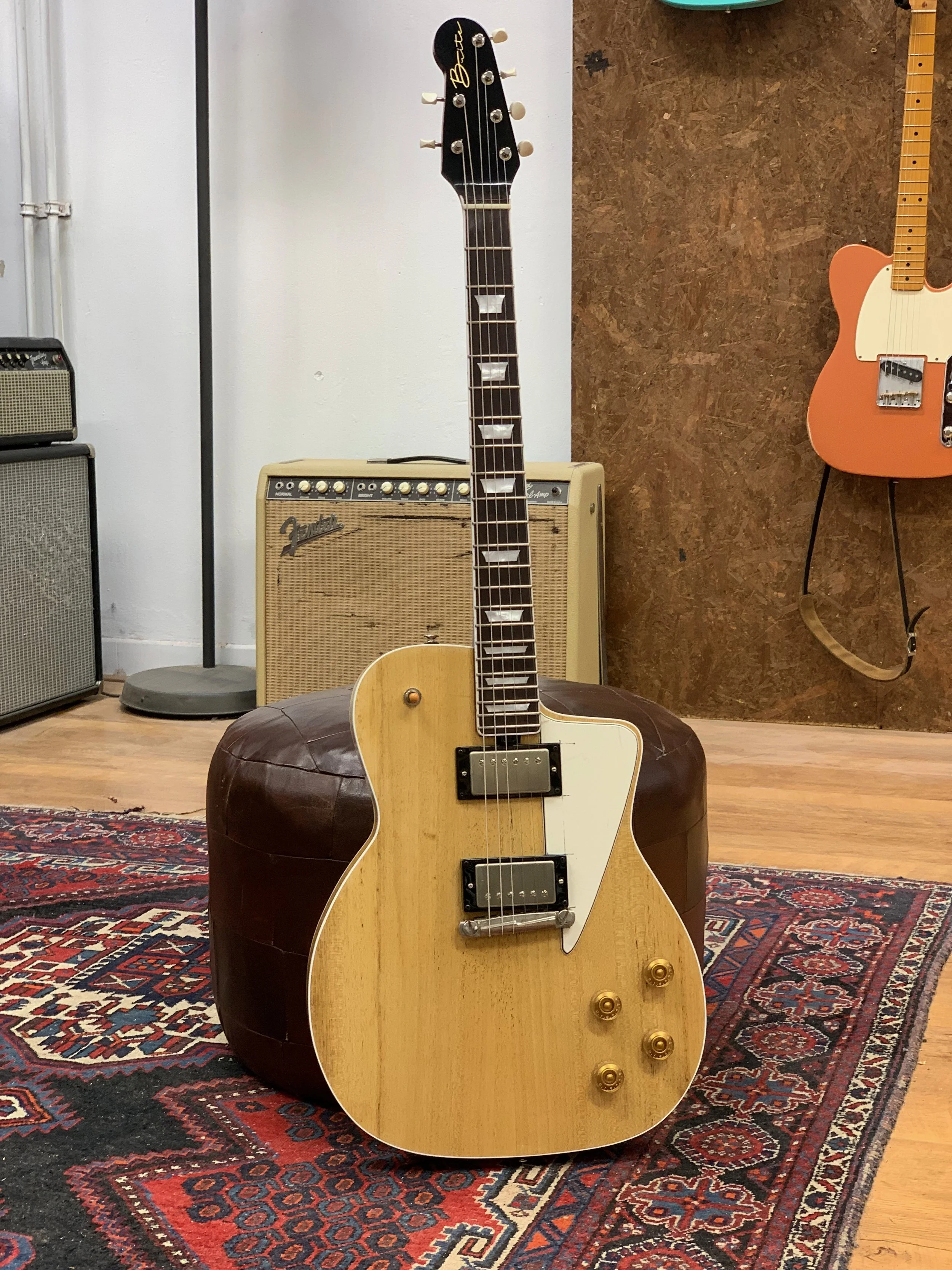 Een akoestische elektrische gitaar met een natuurlijke houtafwerking, liggend op een bronzen poef op een traditionele tapijt, in een muziekkamer met versterkers en een andere elektrische gitaar aan de muur.