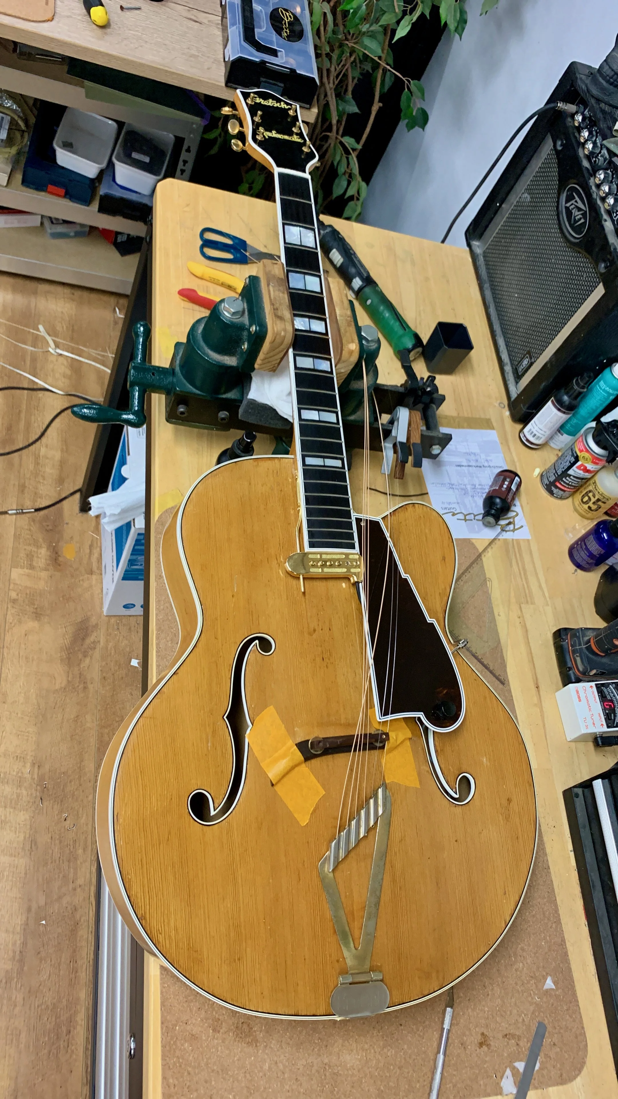 Een houten elektrische gitaar met een gouden tremolo en sierlijke f-holes ligt op een werkbank. De gitaar wordt gerepareerd of onderhouden in een werkplaats, omringd door gereedschap en onderdelen.