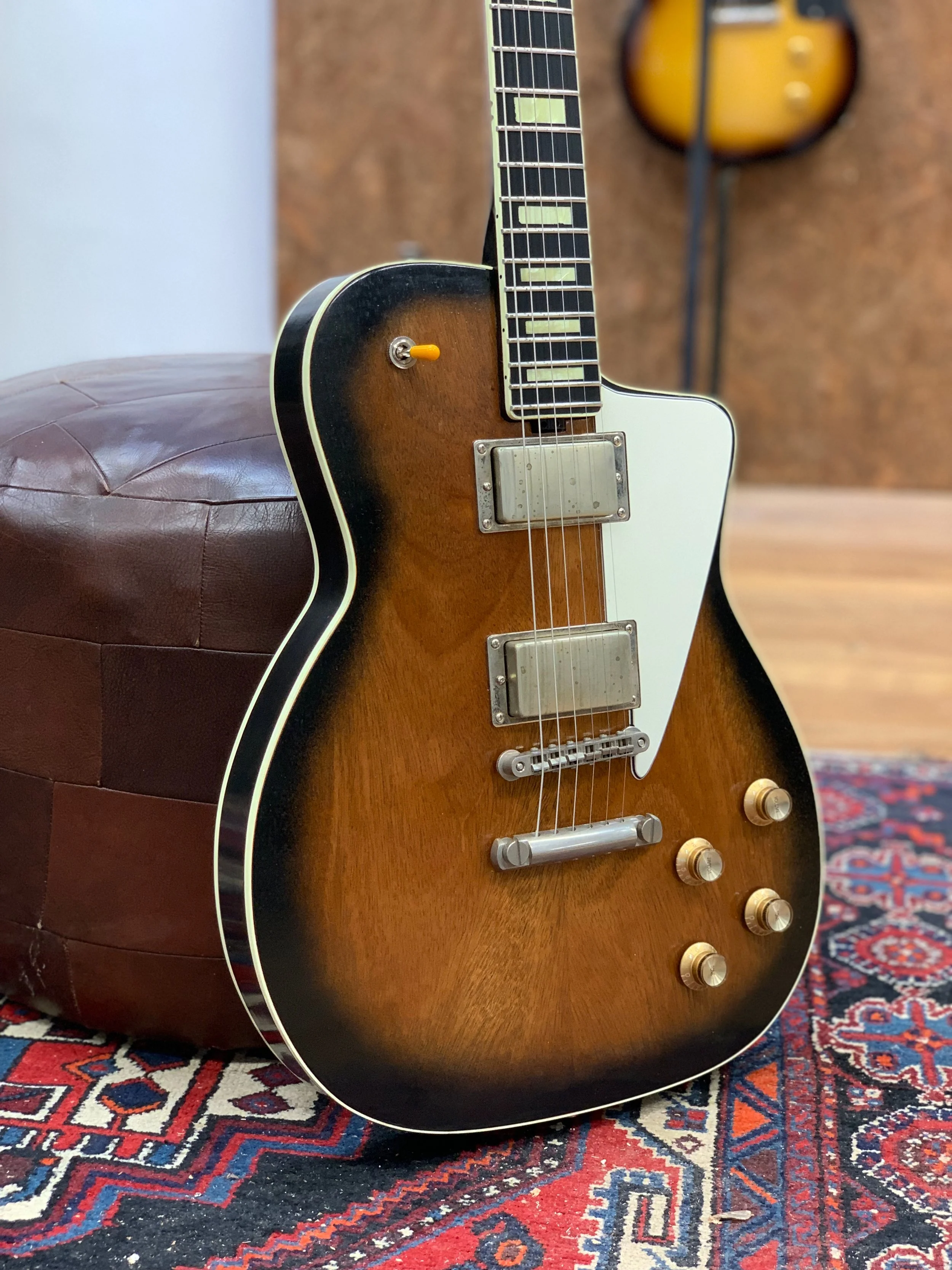 Een vintage elektrische gitaar met een bruin en zwarte body, liggend op een tapijt. Op de muur op de achtergrond hangt een andere gitaarluit en een muur met een houtlook.