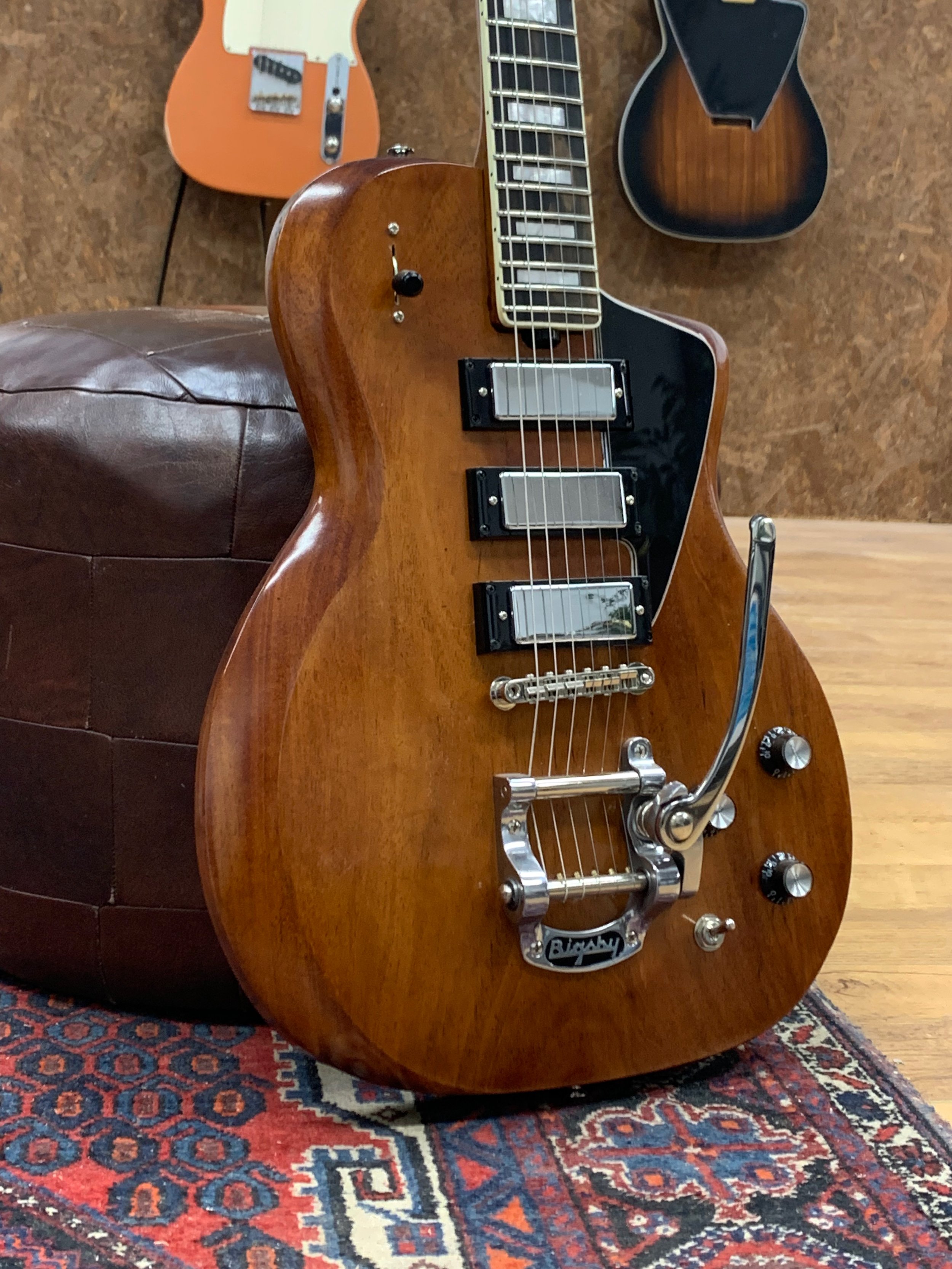 Een houten elektrische gitaar ligt op een tapijt met een patroon, naast een leren armstoel en een andere gitaartas op de vloer.