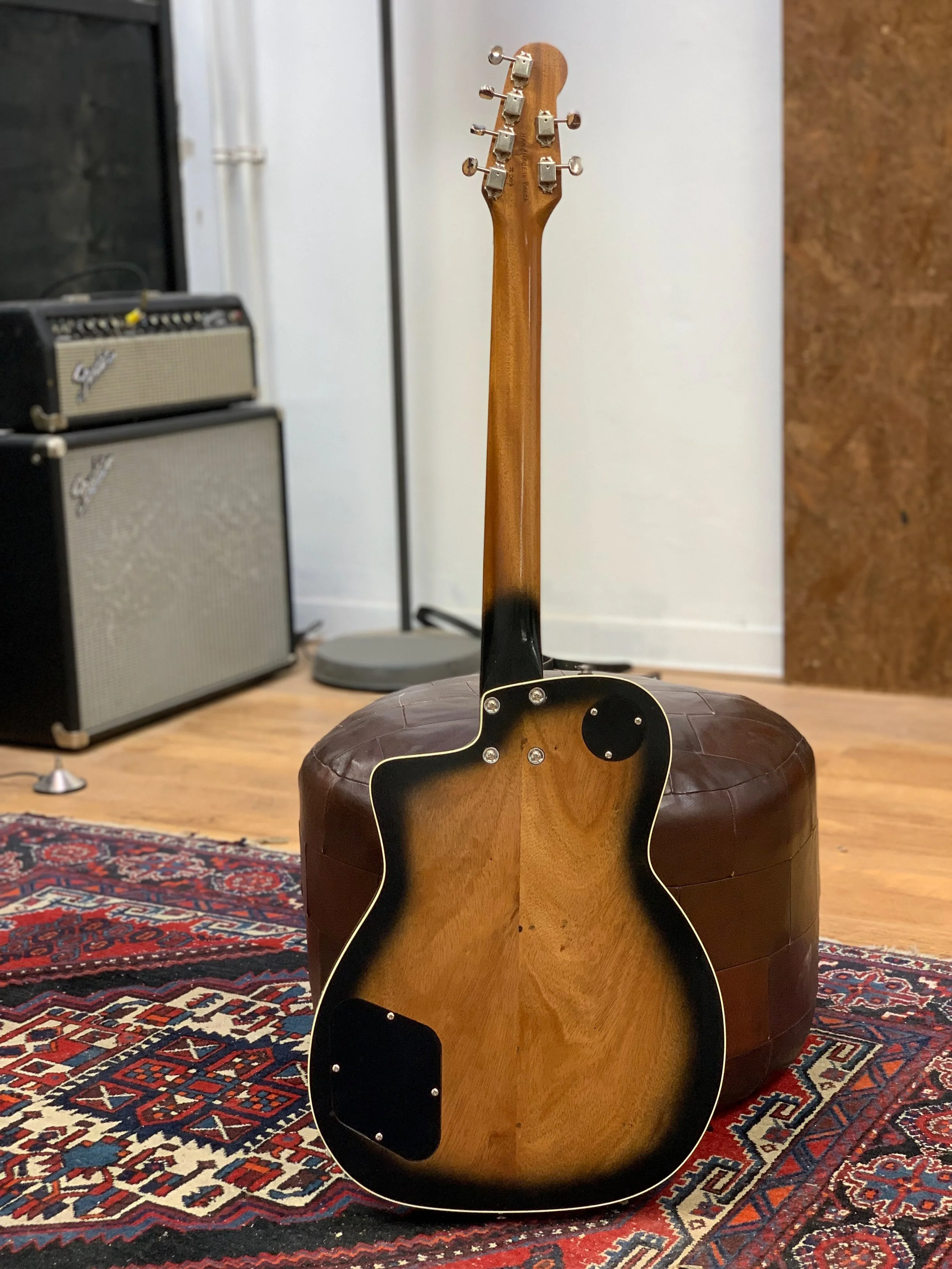 Achteraanzicht van een akoestische gitaar die op een ronde poef staat in een kamer met een Perzisch tapijt op de houten vloer, met enkele versterkers links en een witte muur op de achtergrond.