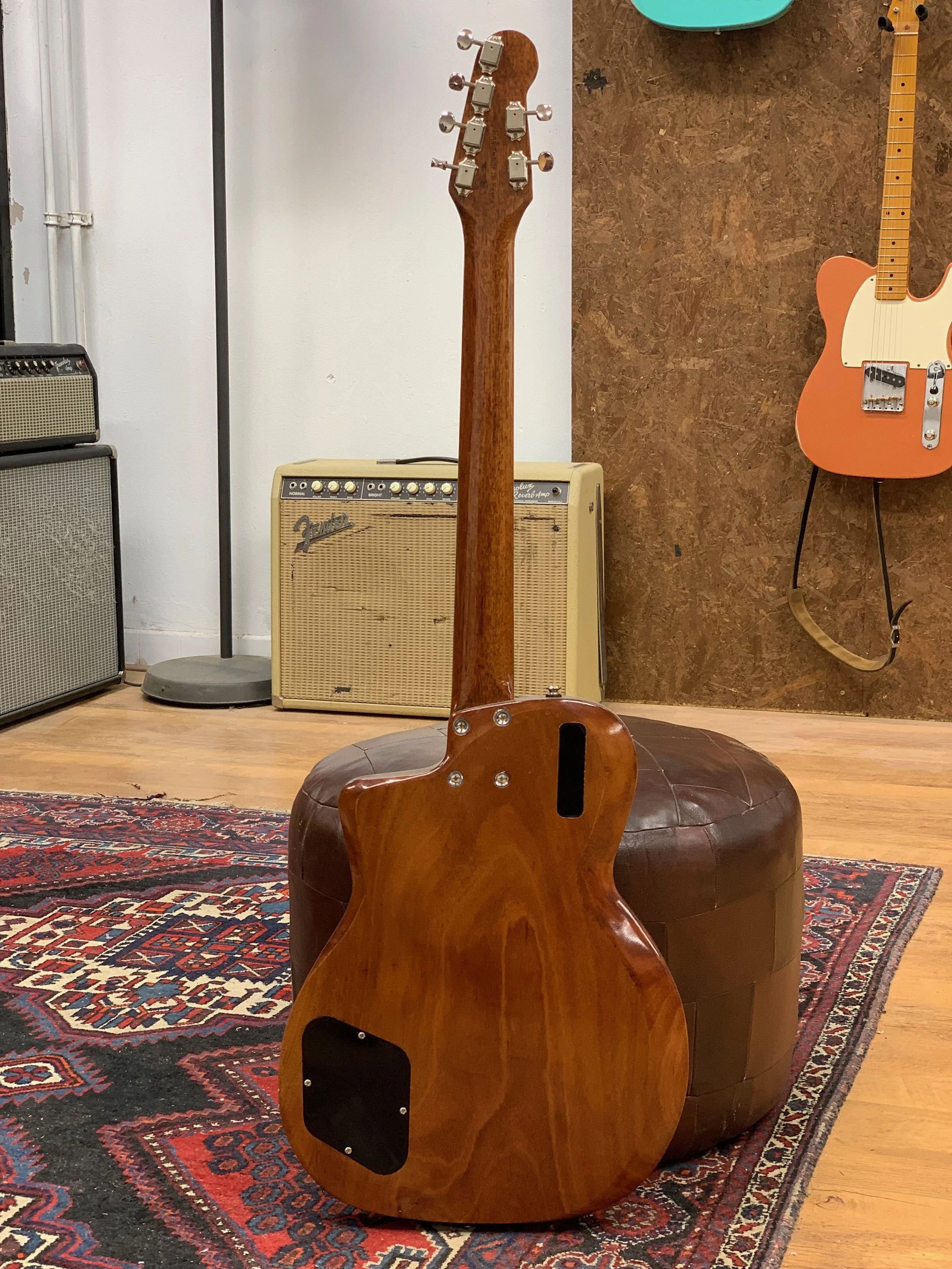 Een elektrische gitaar met een houten achterkant staat op een ronde, geschoten poef. Op de achtergrond zijn een gitaar, een versterker, een vloerlamp en een houten muur zichtbaar, naast een kleurrijk tapijt.