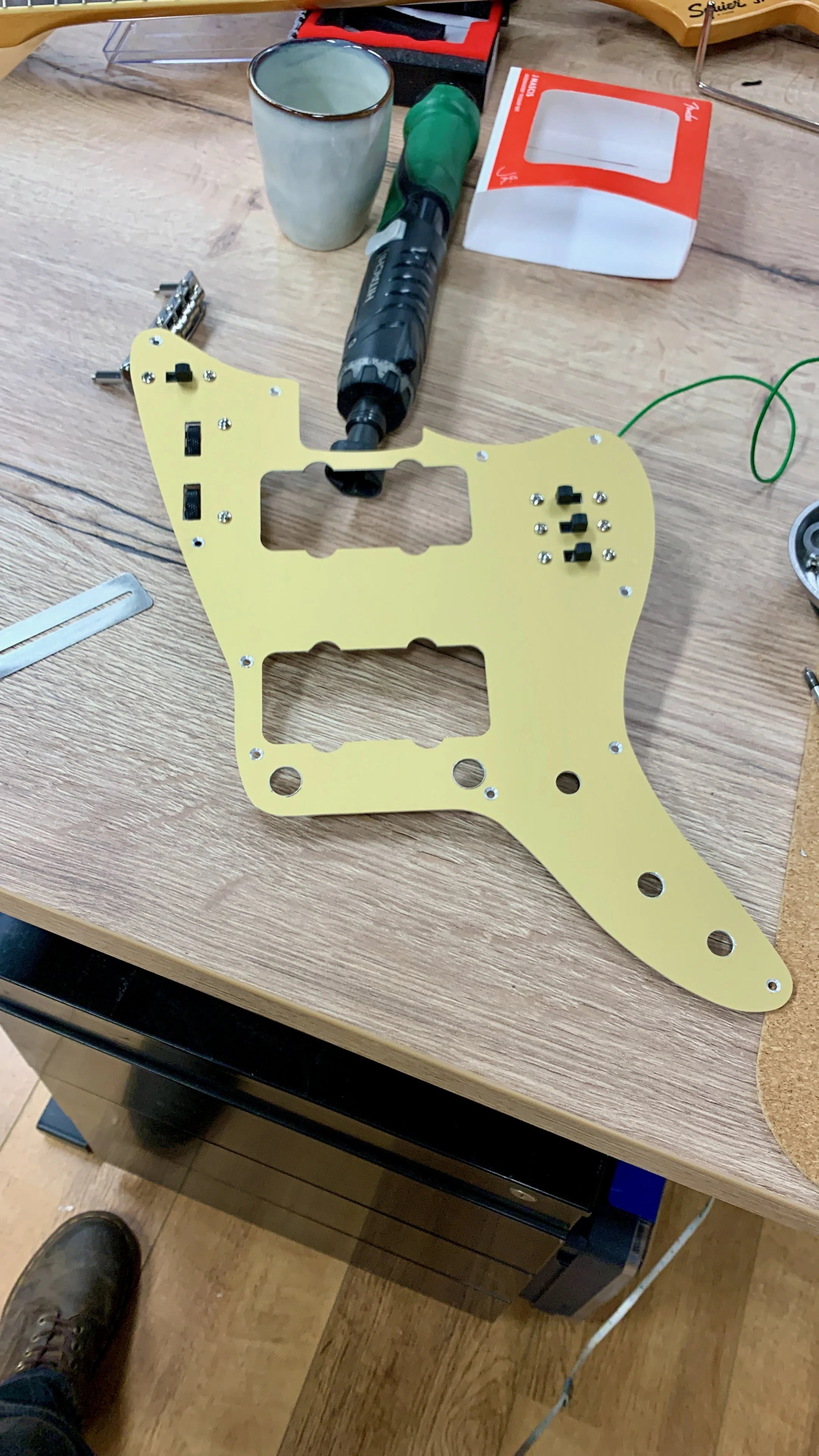 Een houten werkbank met een gele gitaarmontageplaat, een grijs elektrisch gereedschap, een kopje, een doosje met een doorzichtige raampje, een liniaal en een andere gereedschap.