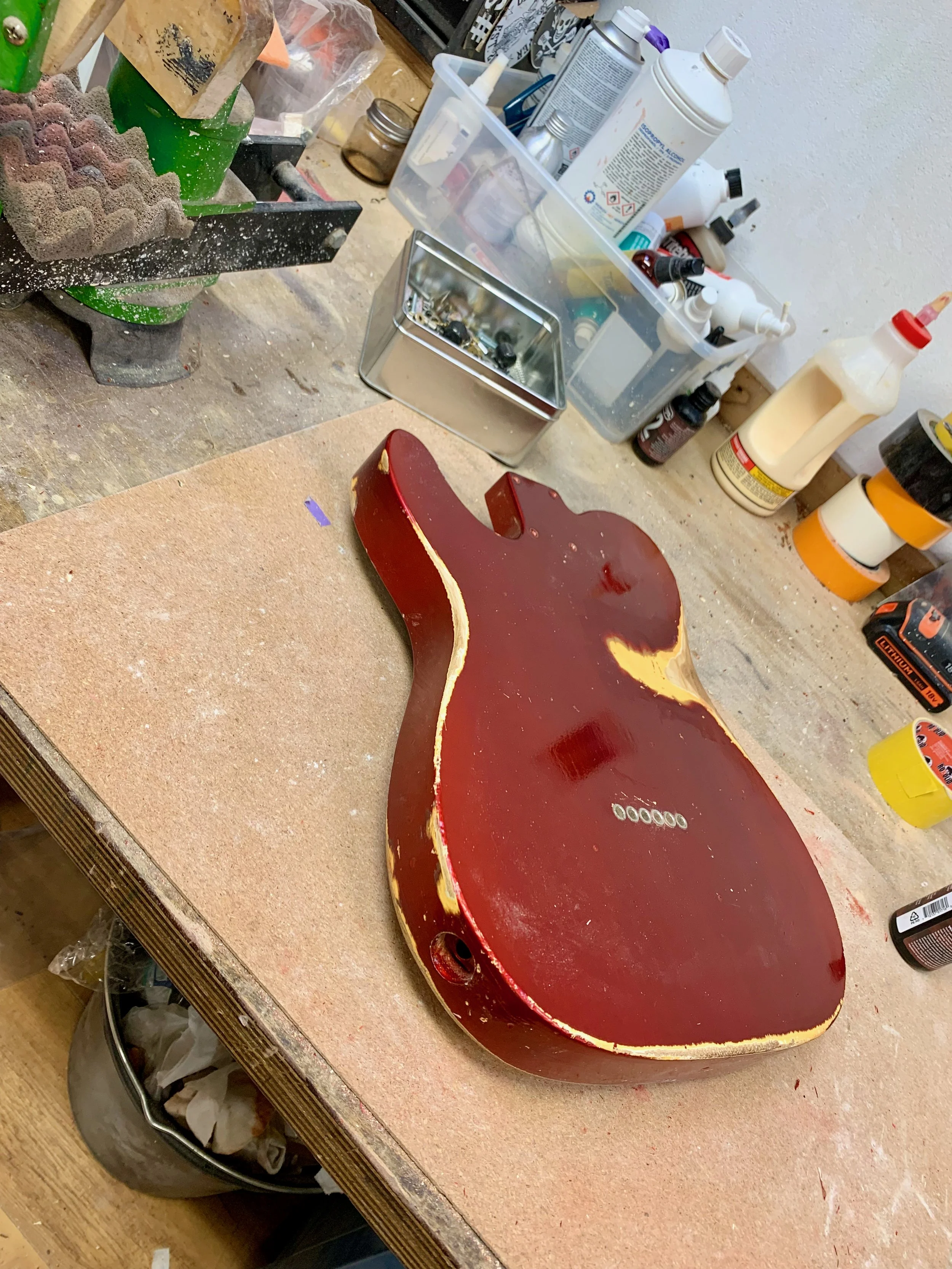 Een werkbank met gereedschap, verf, lijm en materialen, en een bijna afgewerkte elektrische gitaar zonder snaren, met een rode body en houtkleurige rand.