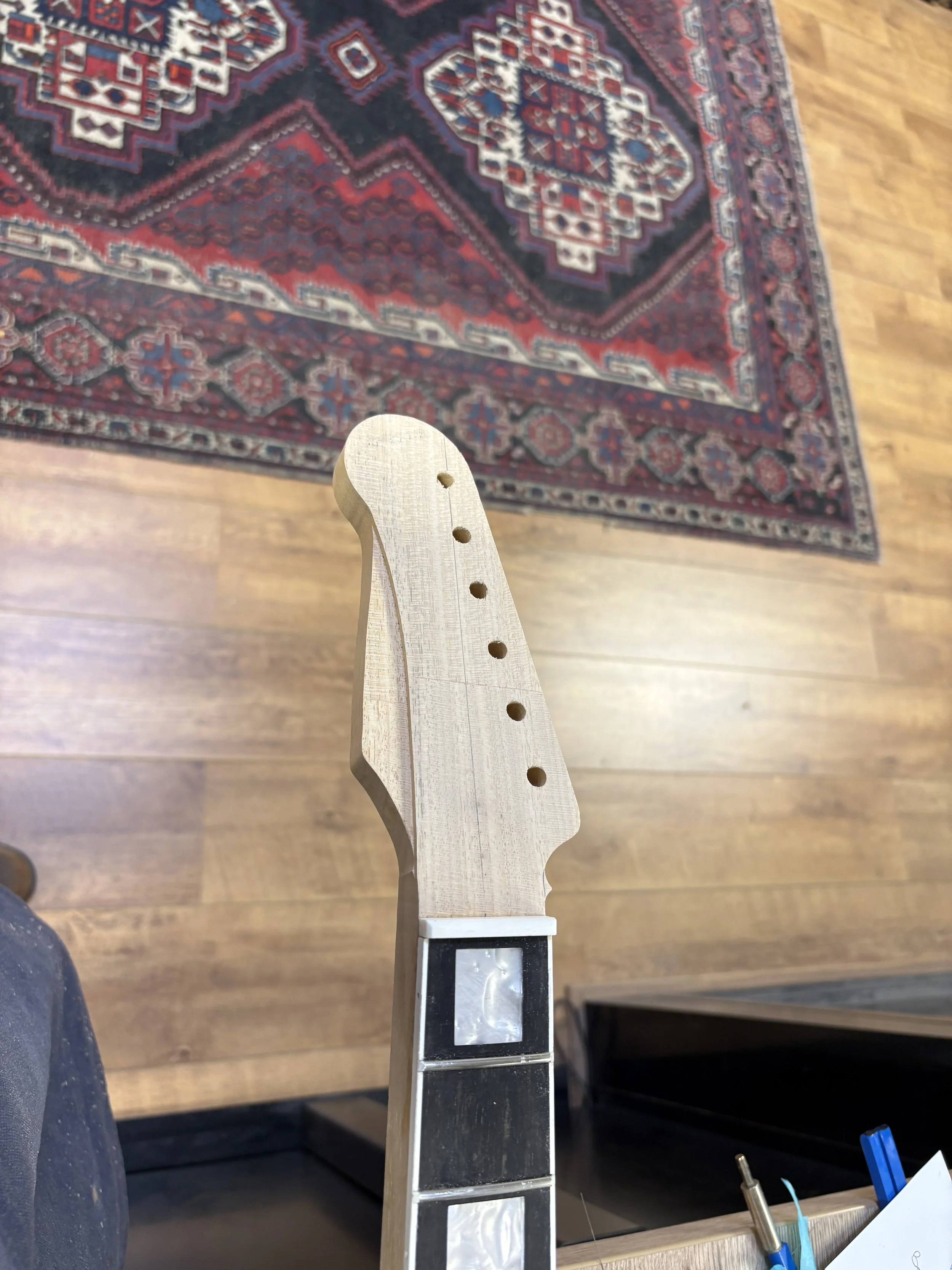 Een onvoltooide gitaarhals van hout op een werkbank in een werkplaats, met gereedschap en een tapijt op de vloer.