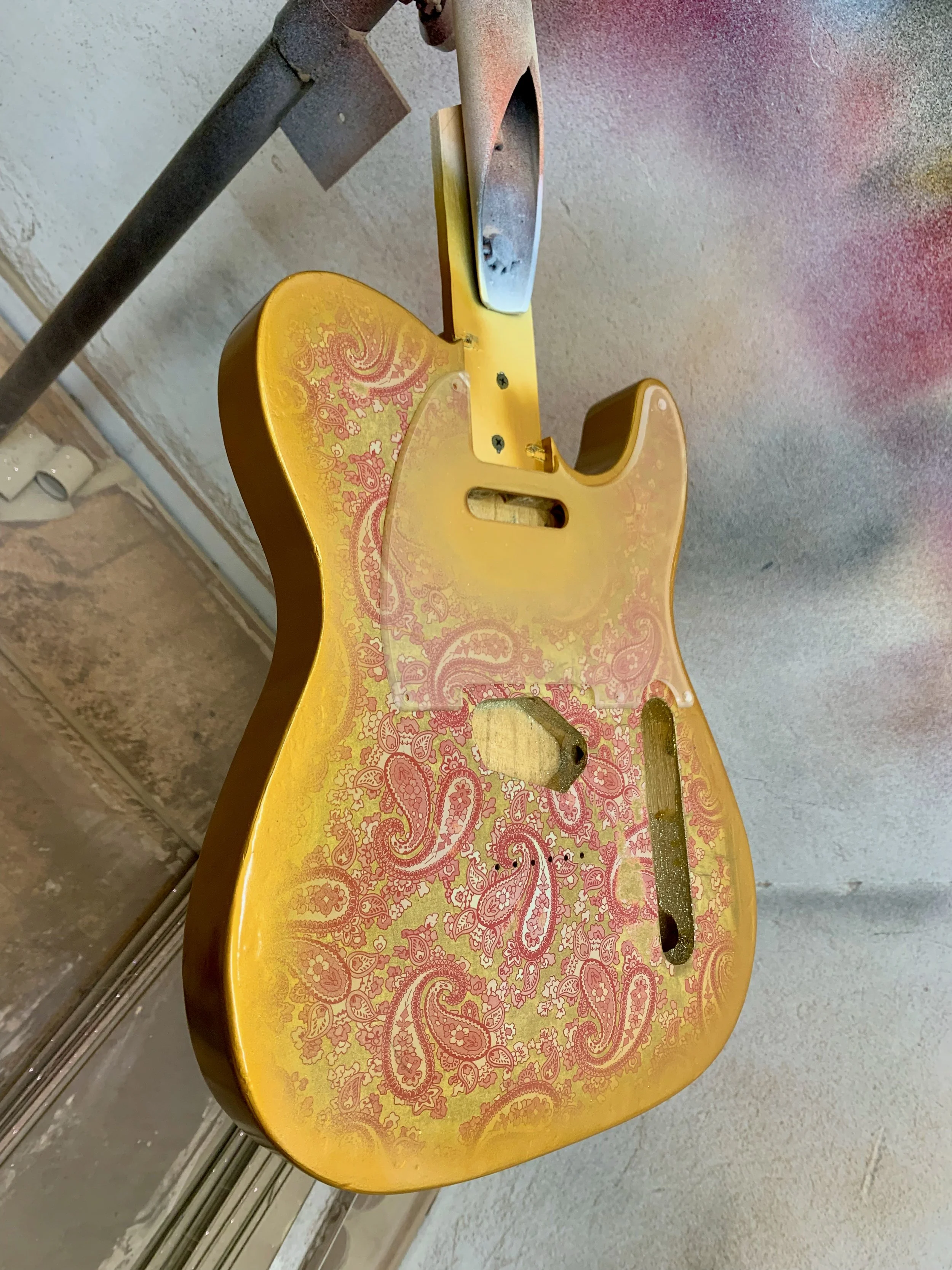 Een geel elektrische gitaar wordt zonder snaren en met een onvolledige body op een tafel gelegd. De body heeft een roze patroon met paisley-ontwerp.