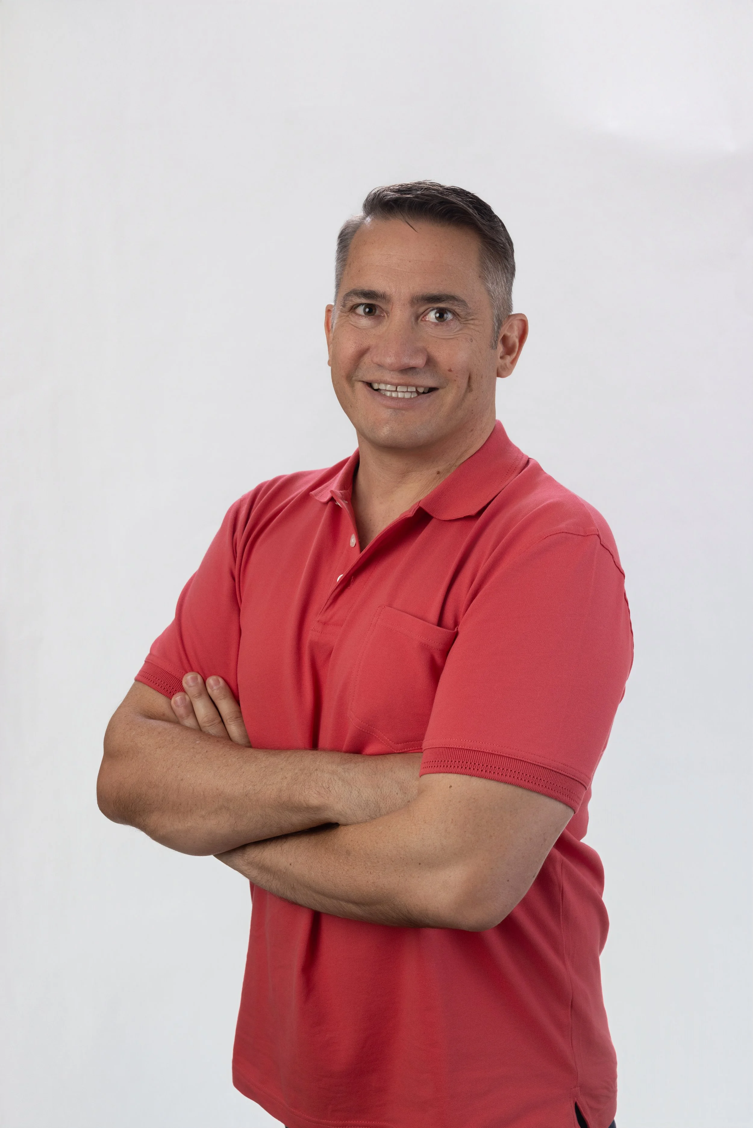 A man with short dark hair, smiling, wearing a red polo shirt with arms crossed, standing against a plain white background.