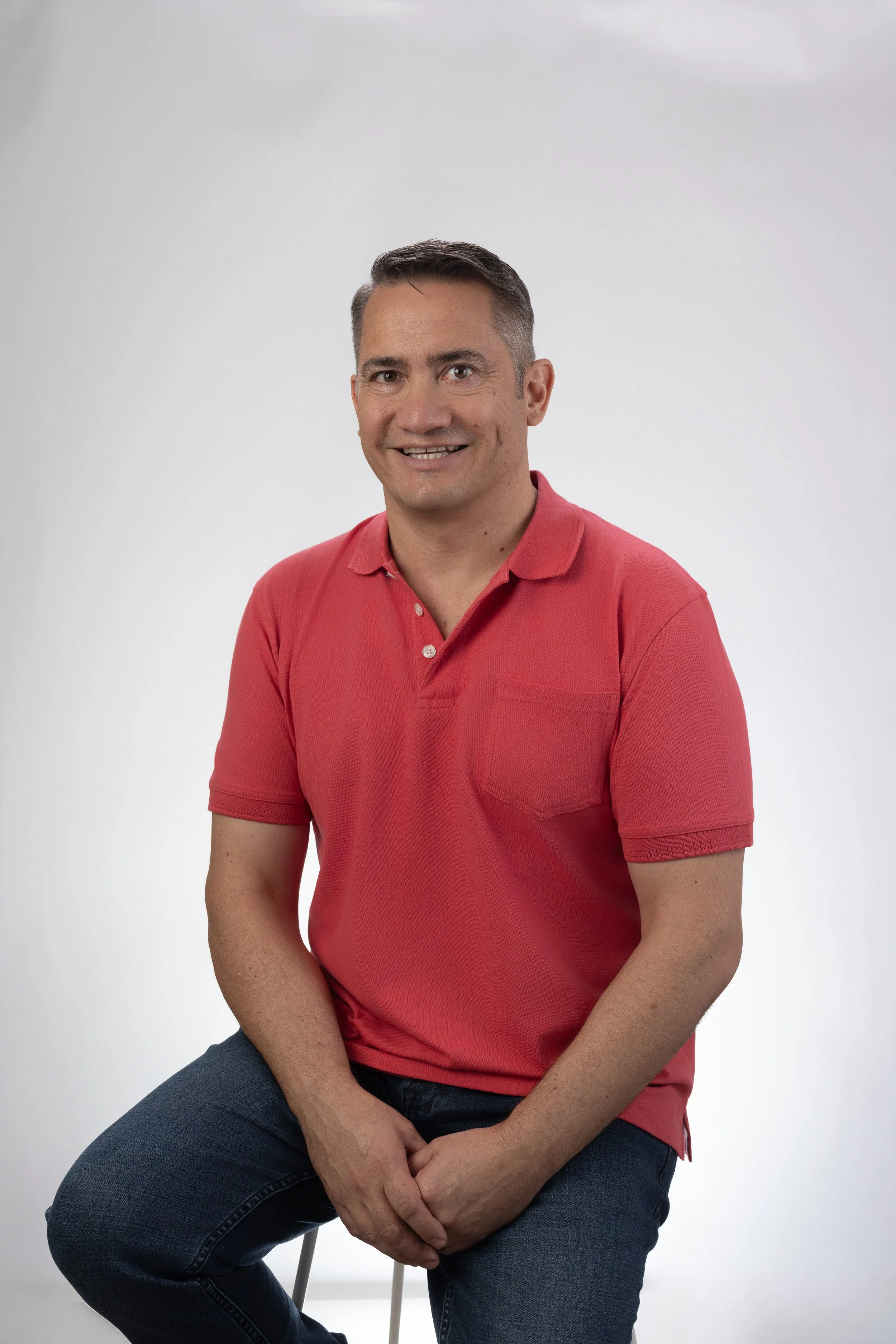 A man sitting on a stool, wearing a red polo shirt and dark jeans, smiling at the camera against a plain white background.