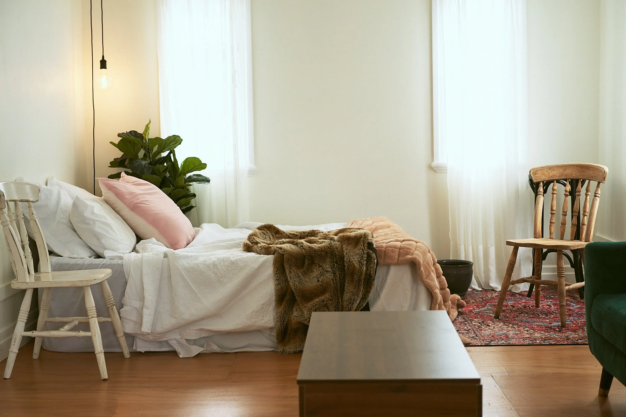 A cozy bedroom with a made bed featuring white sheets, a pink pillow, and a fur blanket. There are two wooden chairs, a green armchair, a potted plant, and a vintage rug near the window with sheer curtains. An exposed light bulb hangs from the ceilin