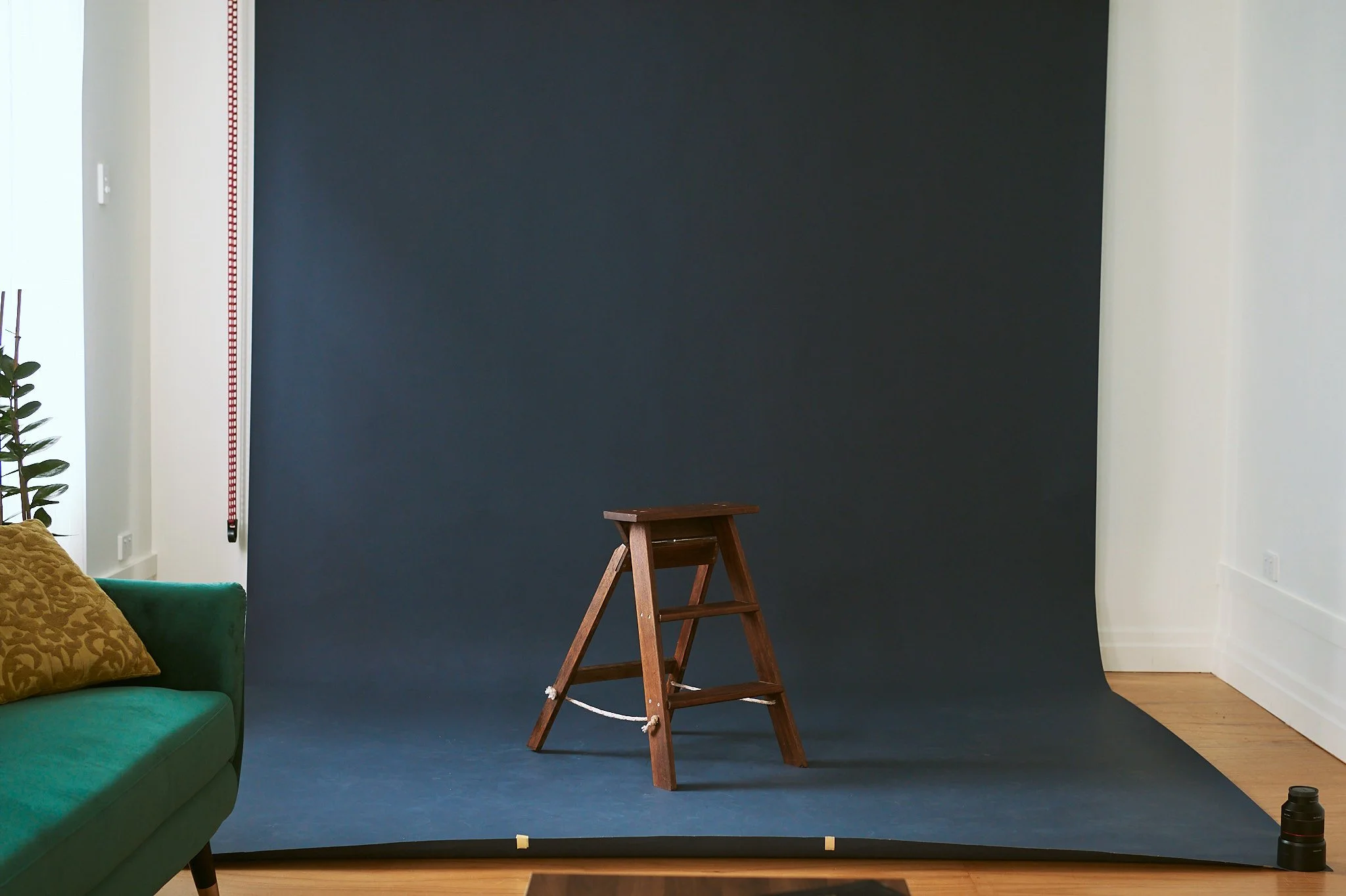 Indoor photography setup with a dark grey backdrop, a wooden step stool at center, a green couch with a decorative pillow on the left, a potted plant, and photography equipment including a camera lens on the right.