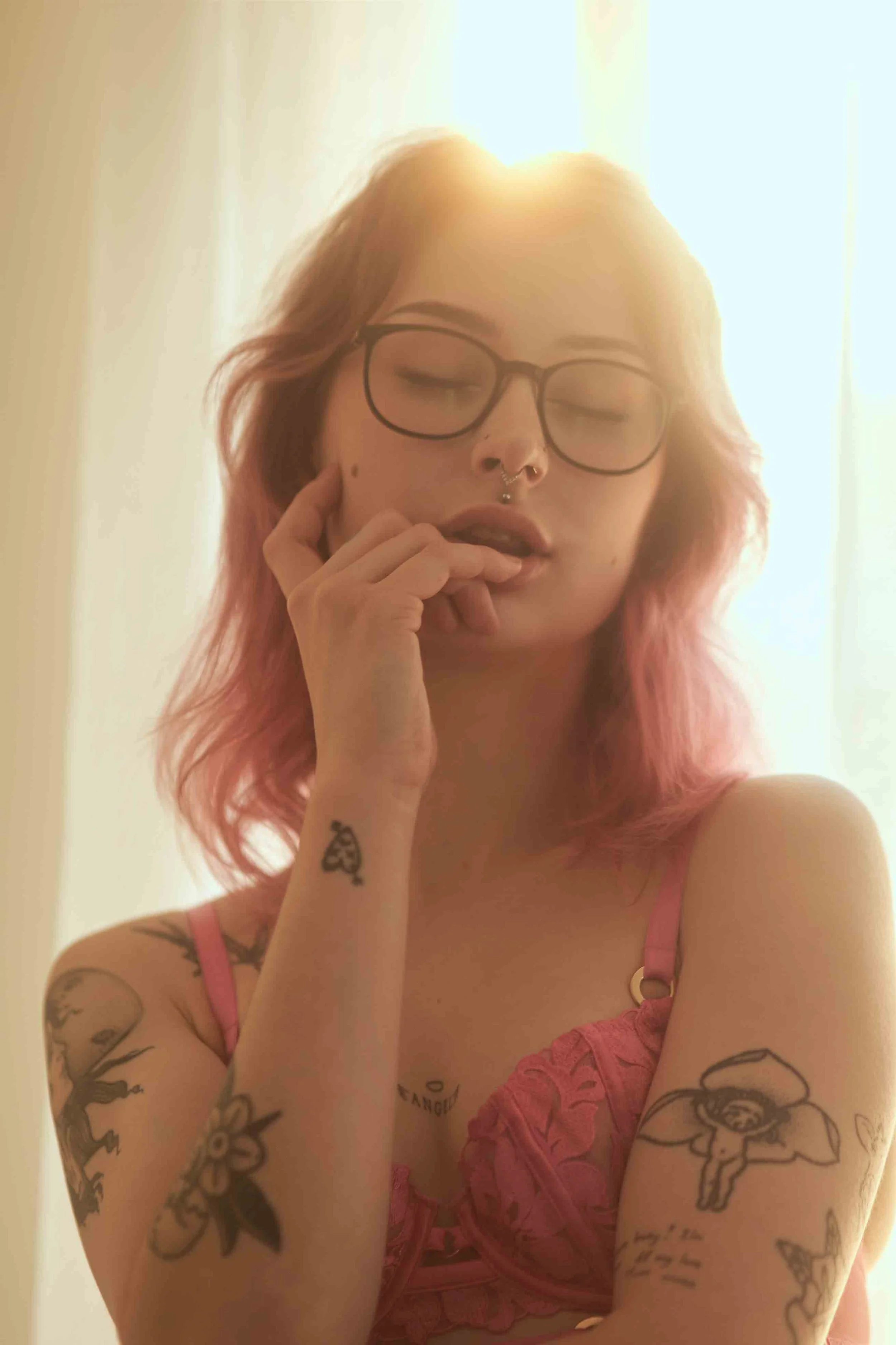 A young woman with pink hair, glasses, and tattoos on her arms, wearing a pink lace top, poses in front of a bright background with sunlight shining behind her.