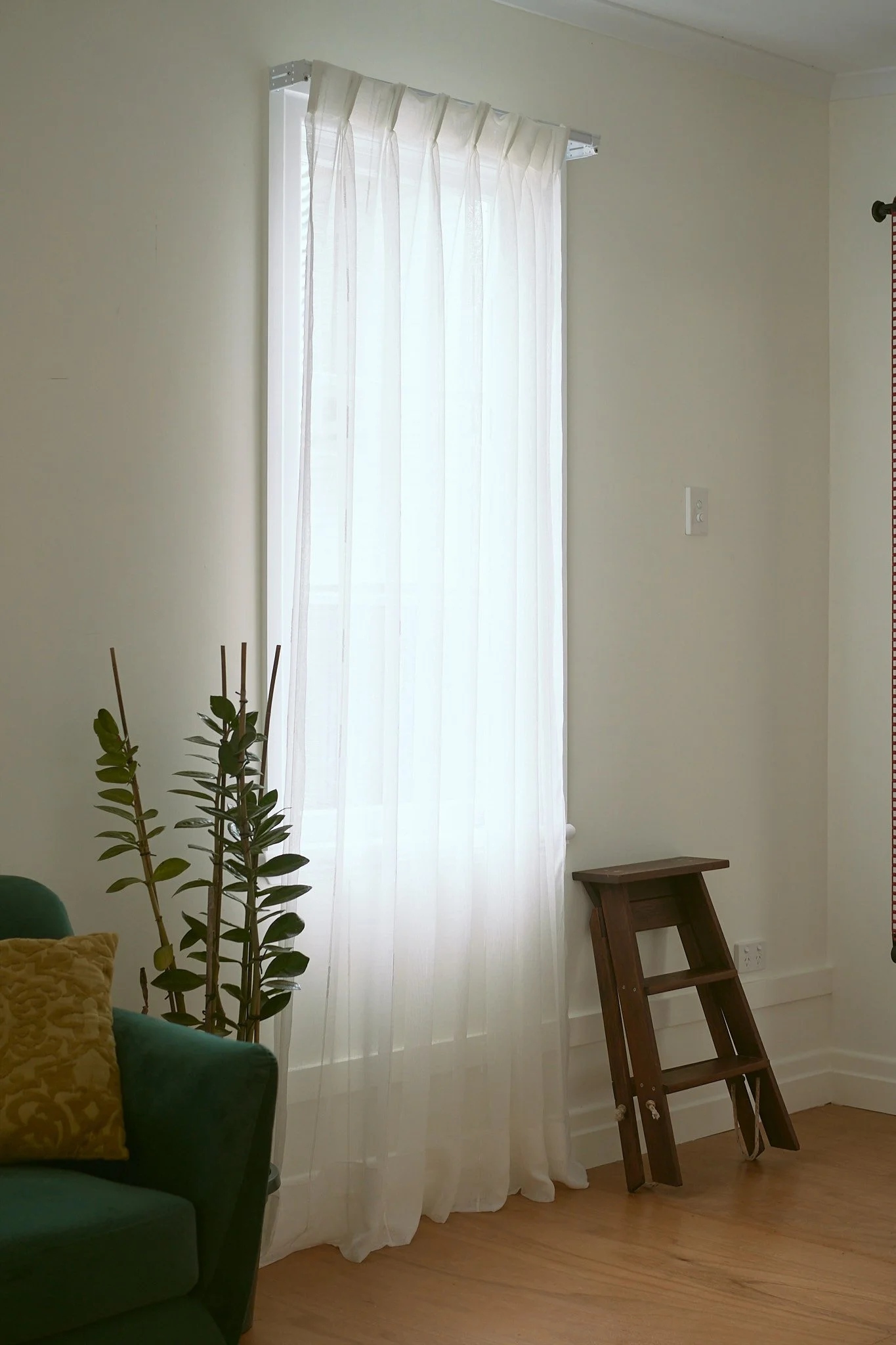 A living room corner with a tall window covered by sheer white curtains. Next to the window is a small wooden step ladder on the light-colored hardwood floor. Part of a green upholstered sofa with a yellow pillow is visible in the lower left corner.