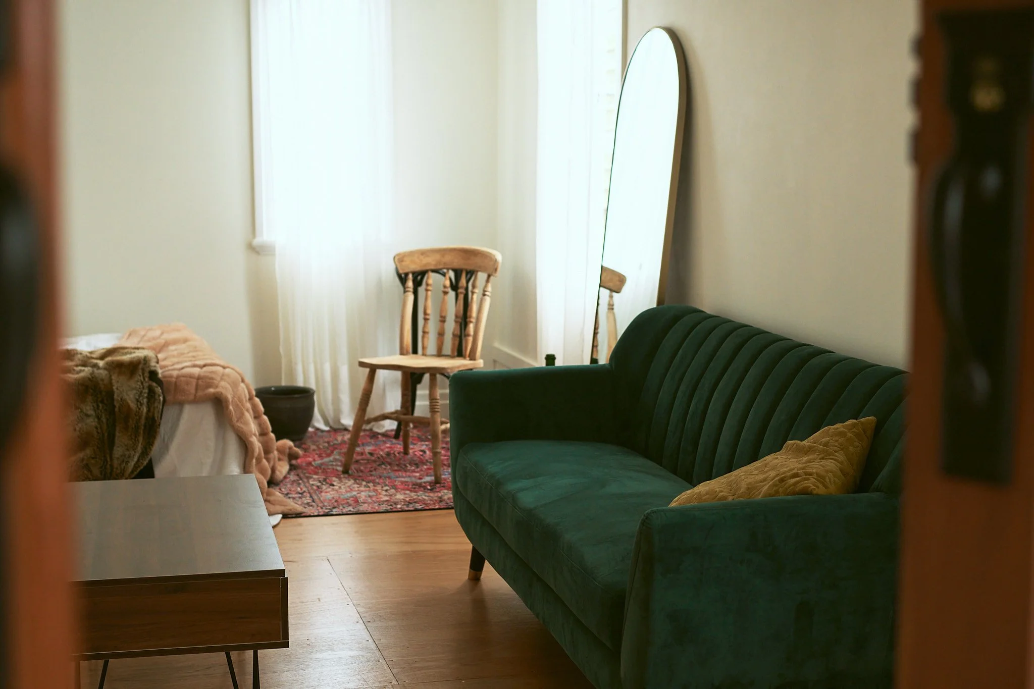 Living room with a green velvet sofa, a wooden chair, a large mirror, a bed with blankets, a small table, and a red area rug, lit by natural light from a window.