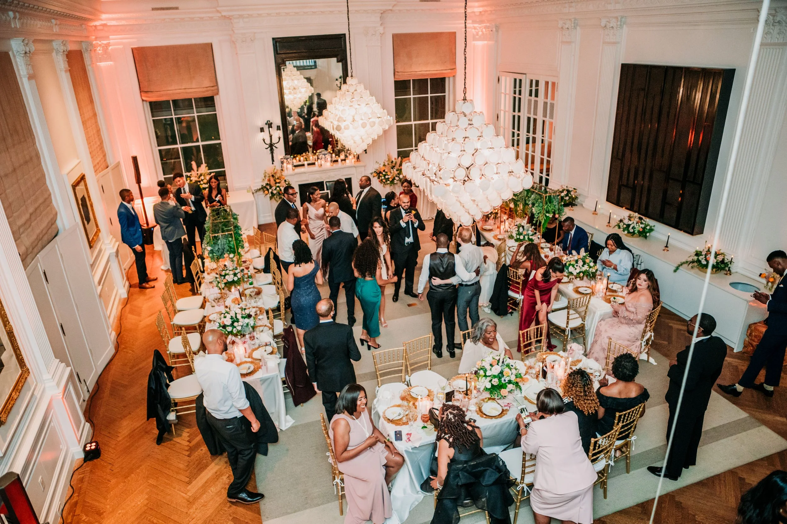Wedding reception with guests dancing and socializing in a large, elegant ballroom decorated with floral arrangements and modern chandeliers.