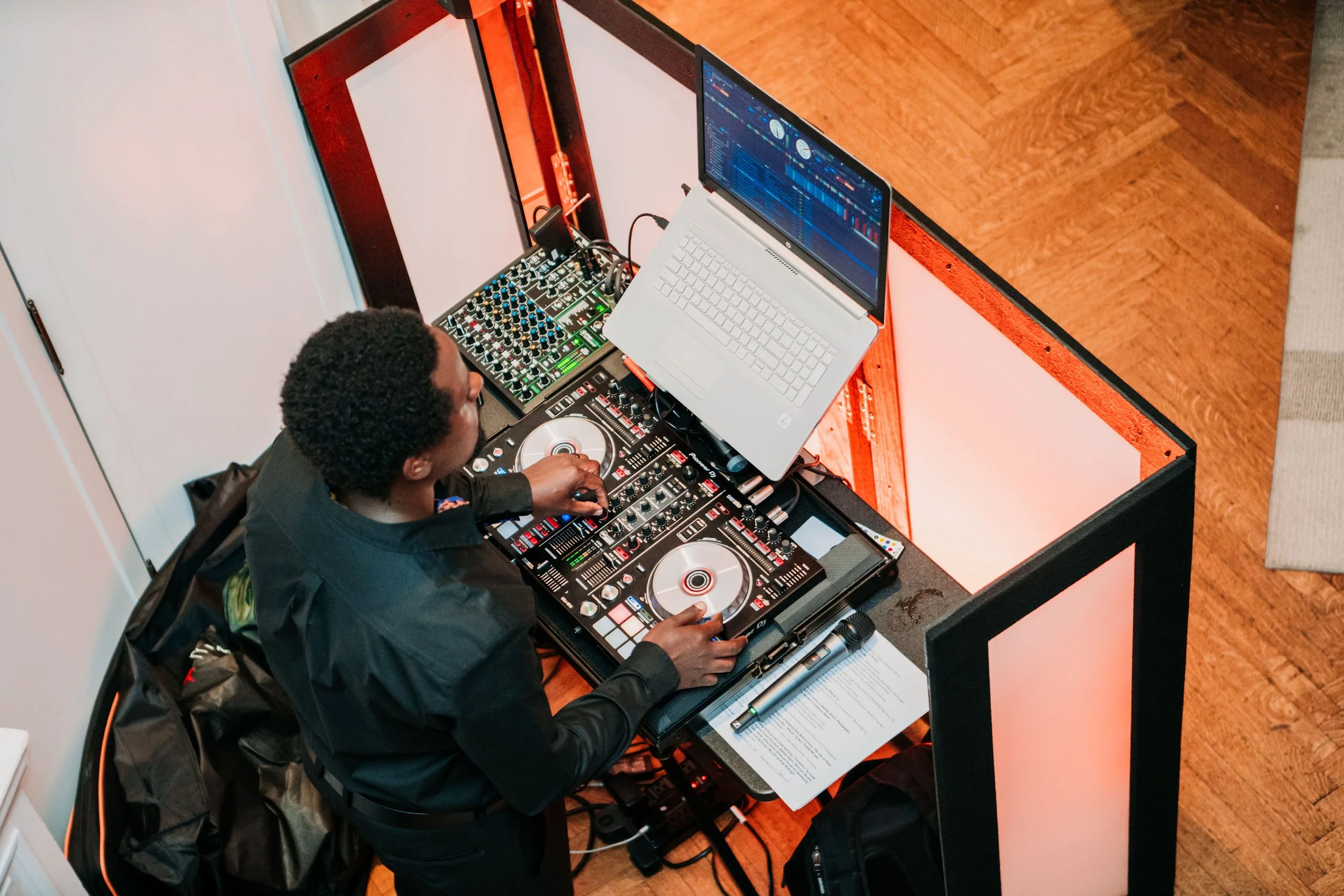DJ mixing music at a booth with turntables, a laptop, and audio equipment, in an indoor venue with wooden flooring.