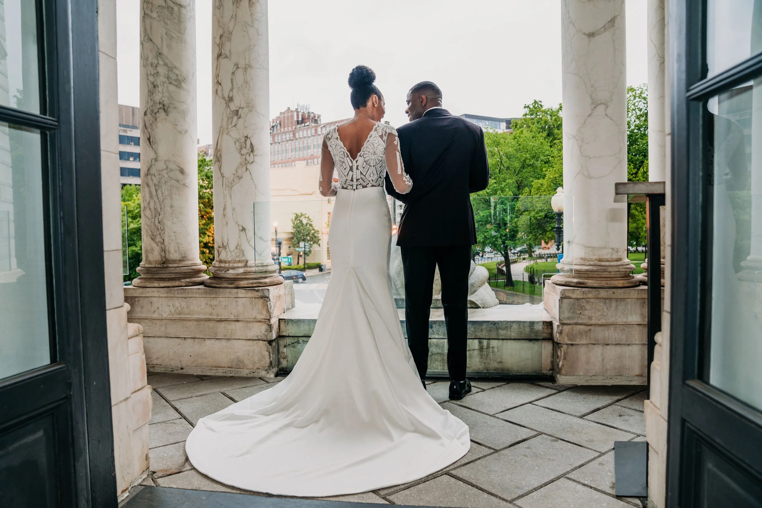 Couple dressed in wedding attire standing on a balcony with marble columns, looking at a city park and buildings in the background.