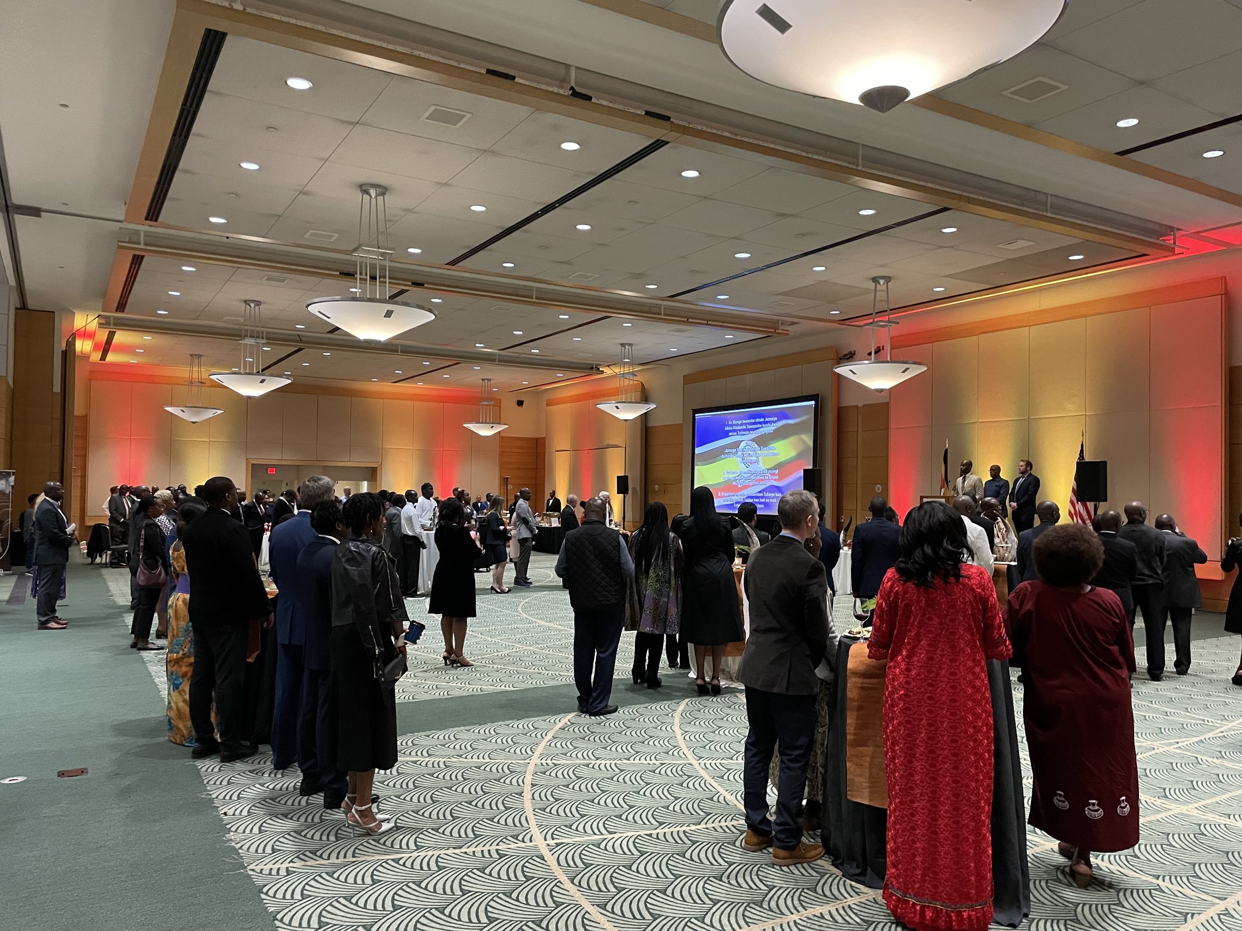 A large group of diverse people attending a formal event in a spacious conference room, facing a stage with a large screen, some standing at high tables, with colorful lighting on the walls.
