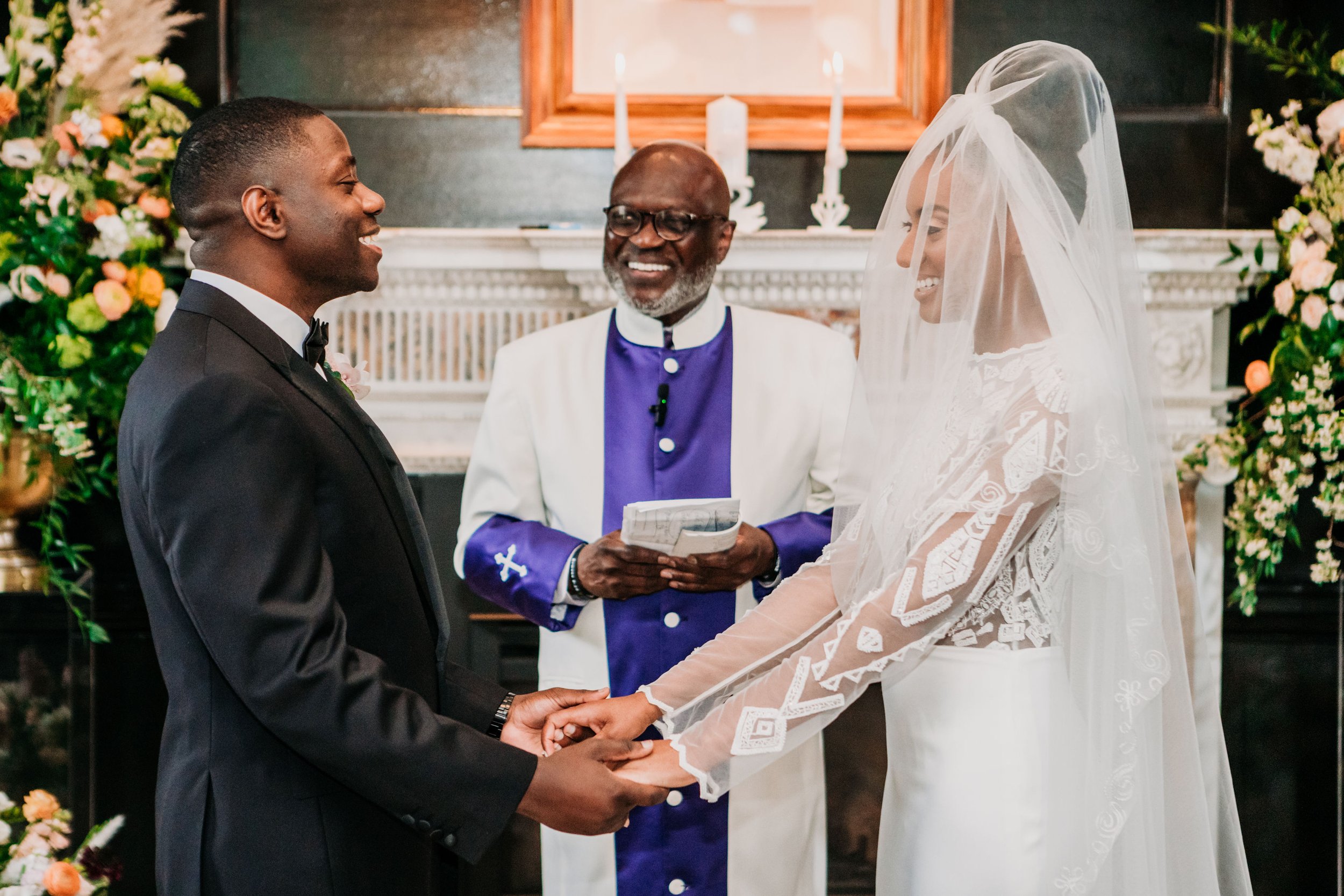 A couple getting married holding hands and smiling at each other, officiant standing behind them with a book in hand, inside a decorated wedding venue with floral arrangements and candles.