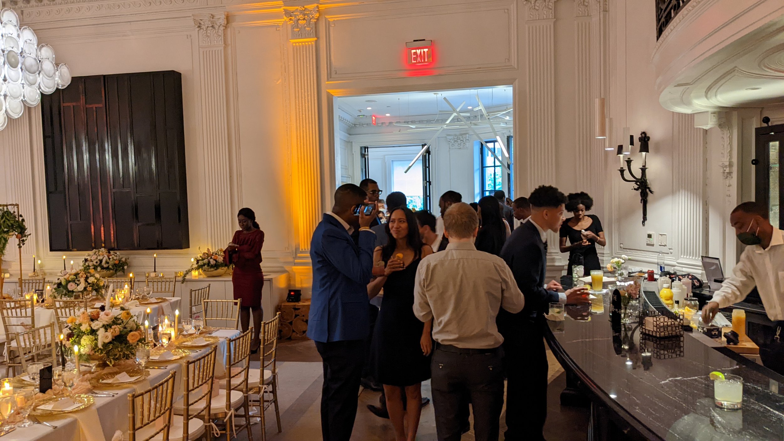 Guests mingling at an elegant event in a decorated hall with floral centerpieces, candlelit tables, and a bar area.