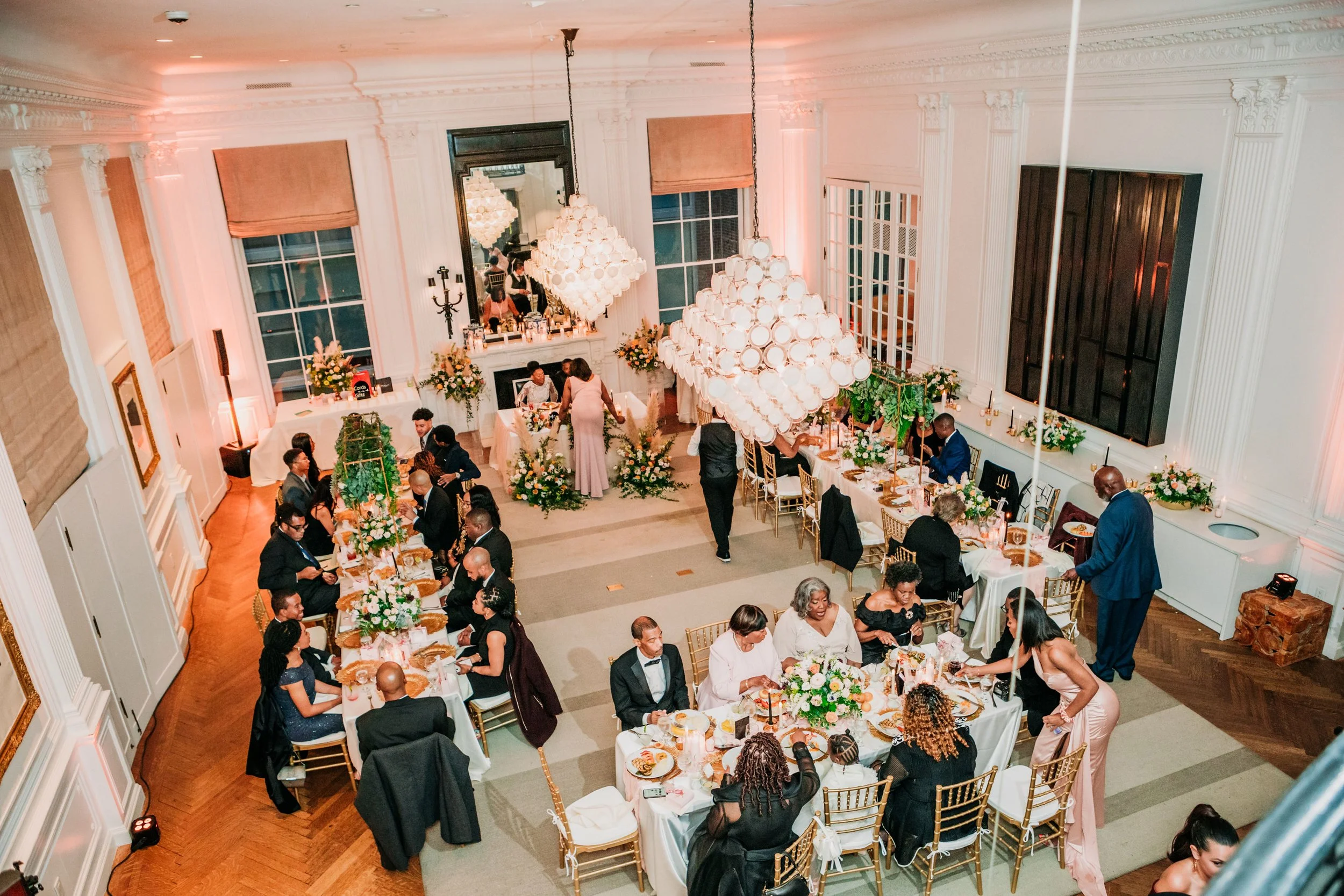 An elegant indoor wedding reception with guests seated at decorated tables, floral arrangements, and large chandeliers hanging from the ceiling.