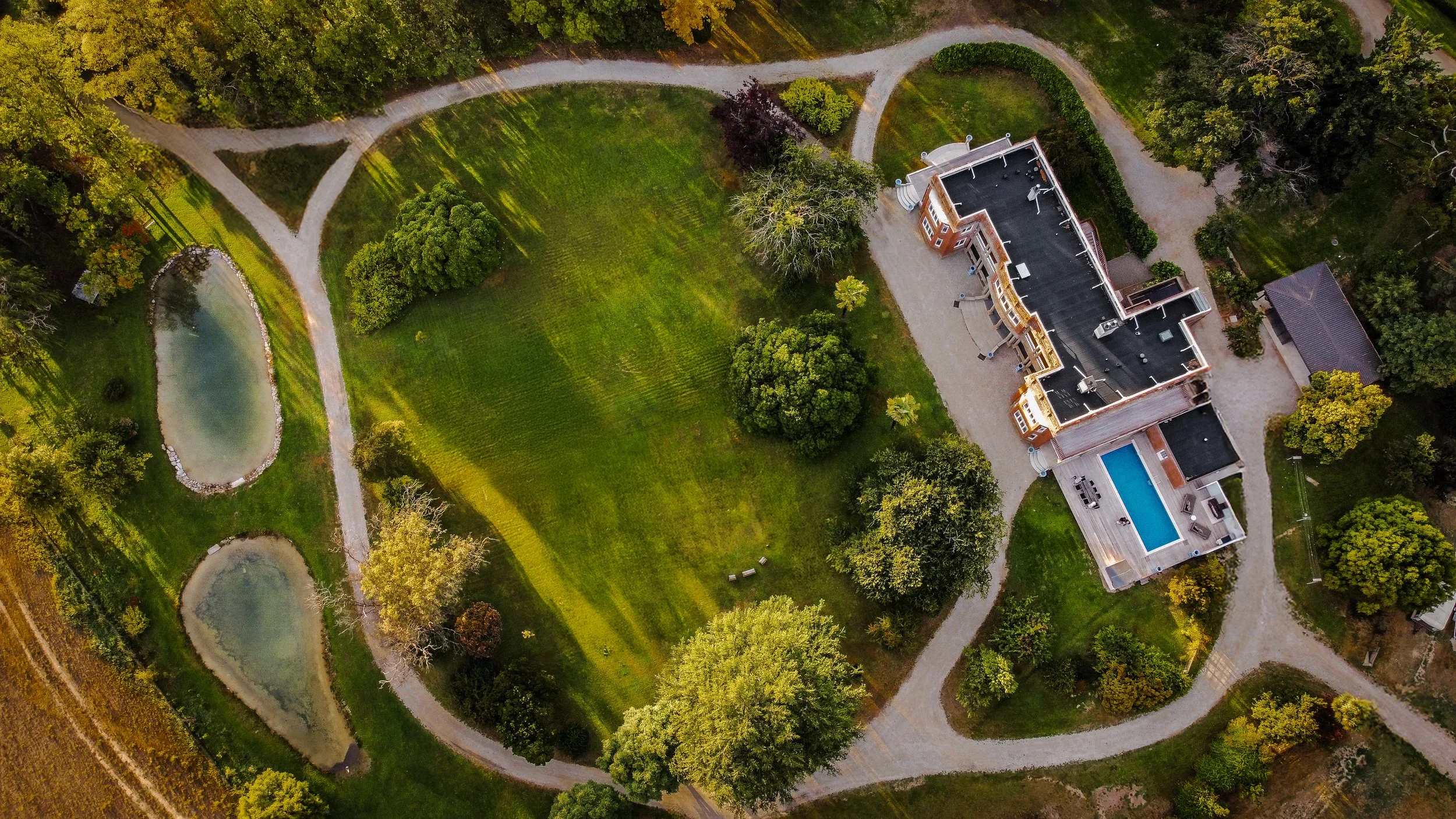 Aerial view of a large estate with green lawns, trees, a swimming pool, a pond, and winding gravel paths surrounded by forest.